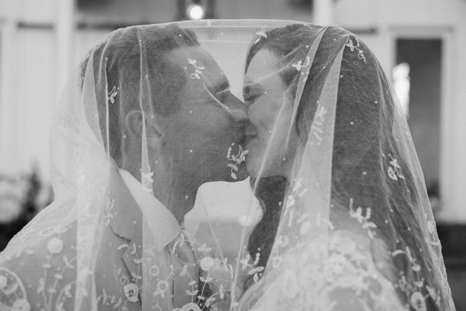 Black and white romantic photo of newlyweds kissing beneath embroidered bridal veil.