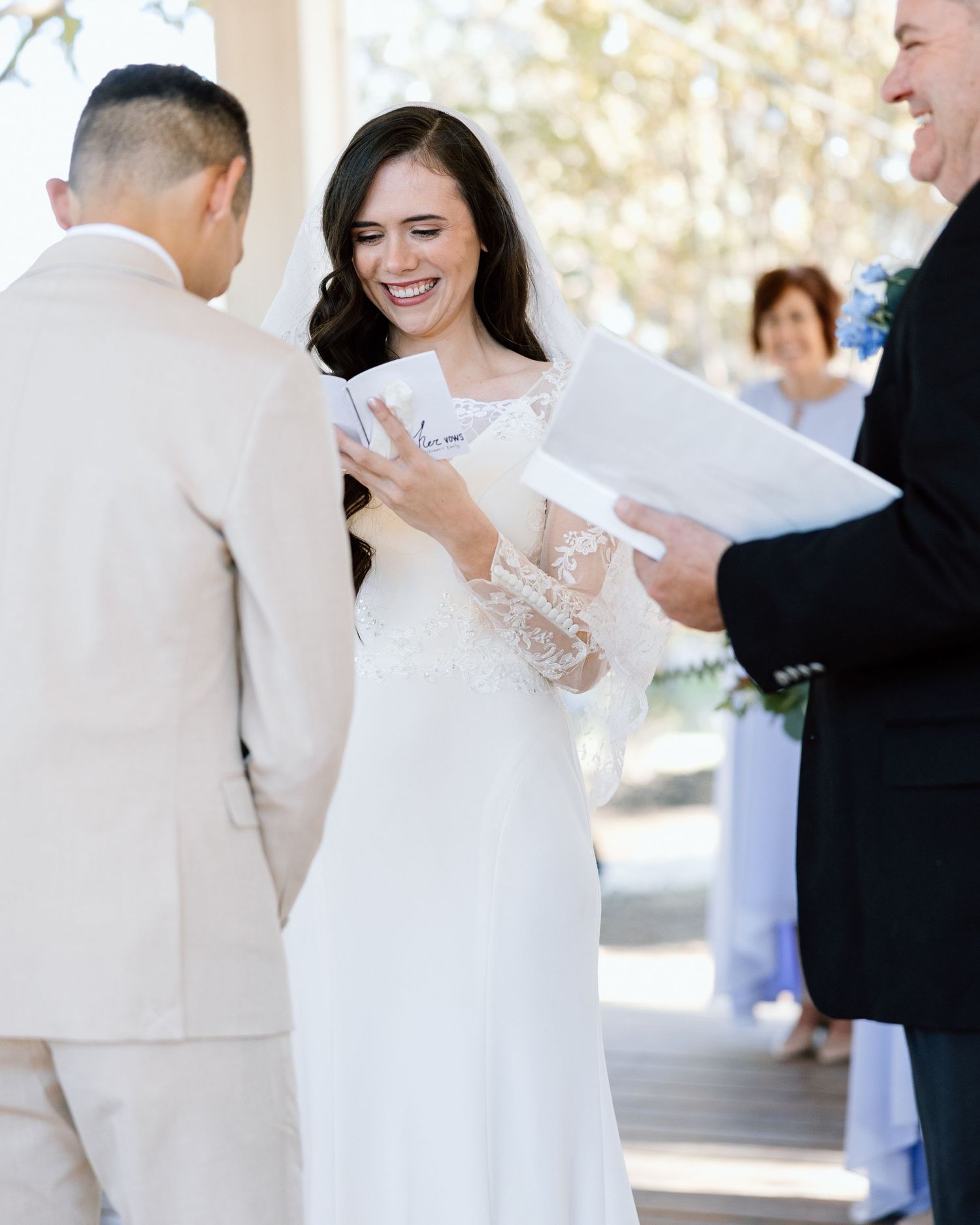 Smiling bride in lace wedding dress reading vows at outdoor ceremony with groom in beige suit.