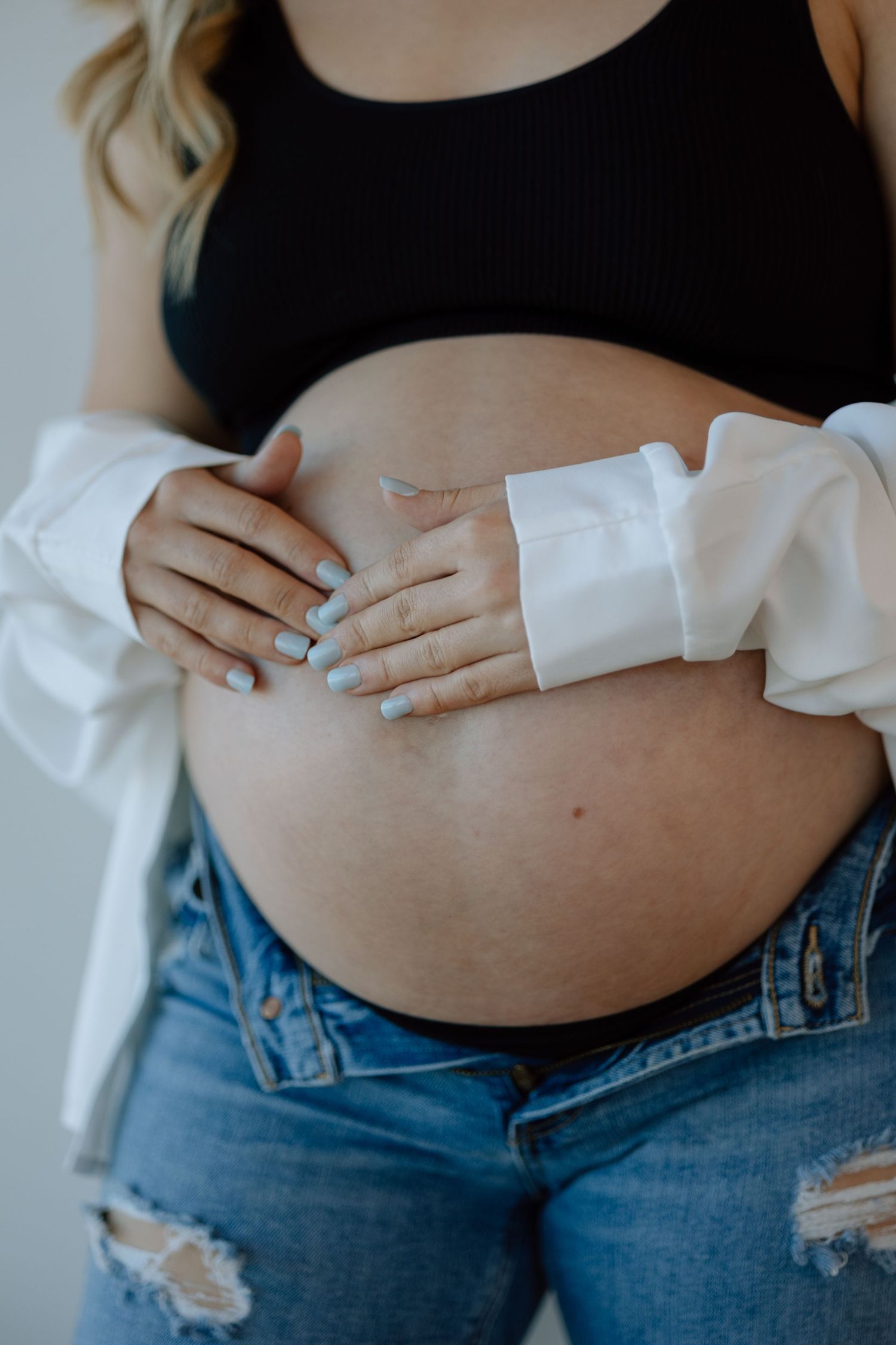 Pregnant woman wearing black top and white cardigan rests hands on bare belly while wearing distressed blue jeans.