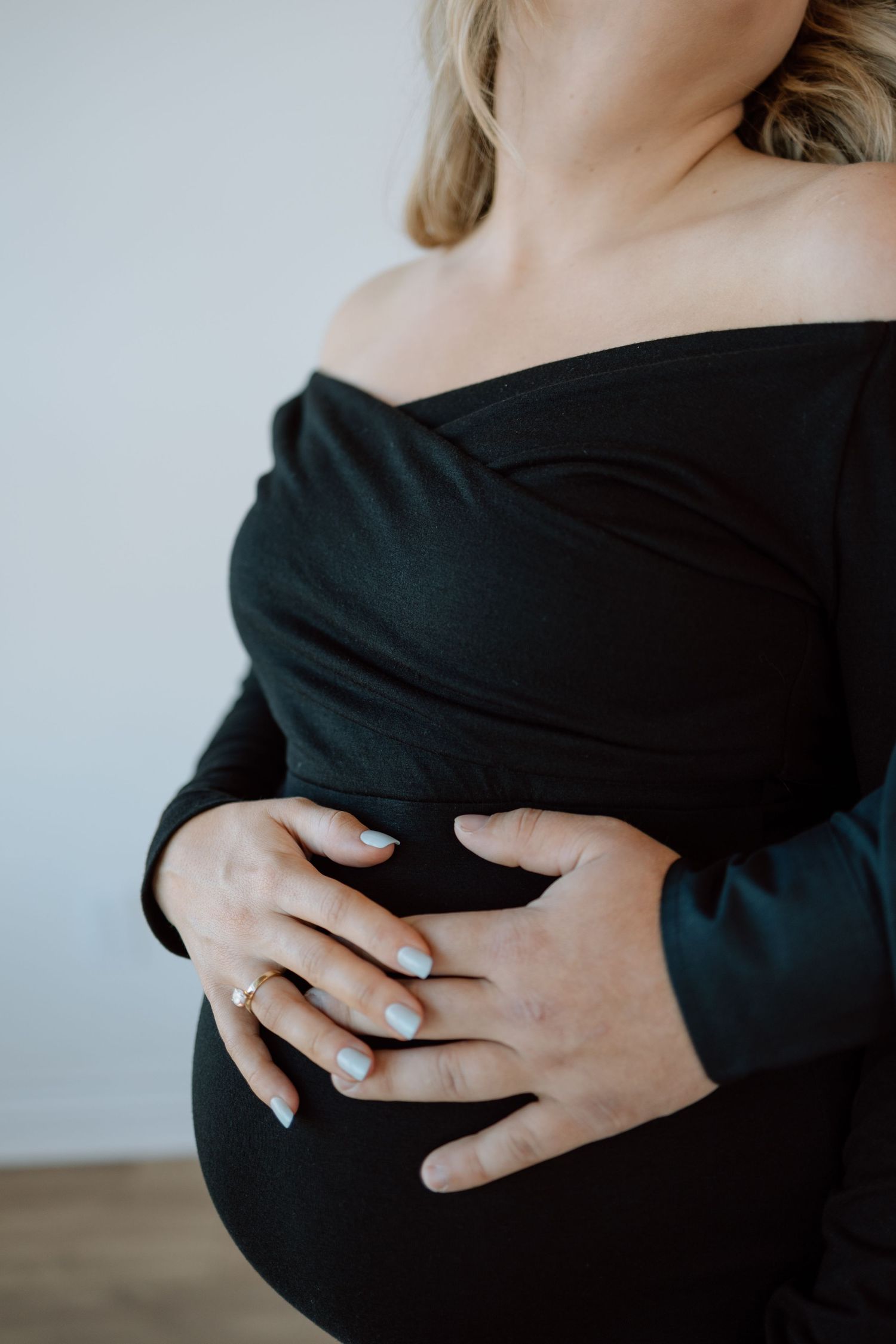 A person in a black off-shoulder top gently cradles their pregnant belly against a light background.
