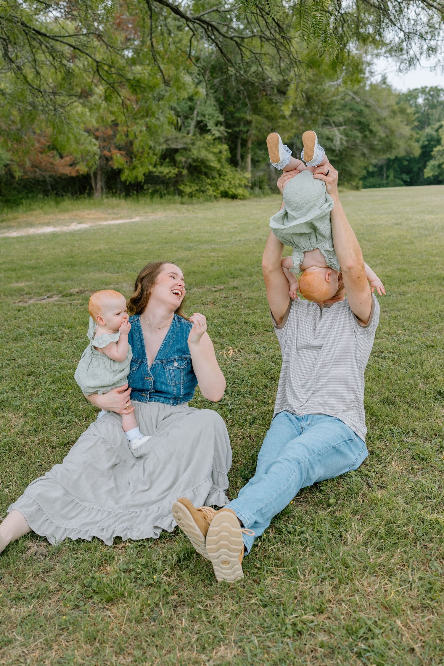 A family enjoys a playful moment together while sitting on grass in a park setting on a sunny day.