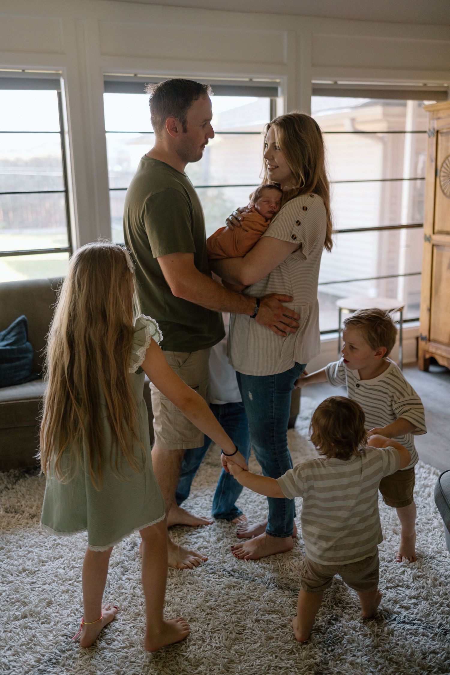 A family shares an intimate moment together in their living room on a gray carpet near windows.