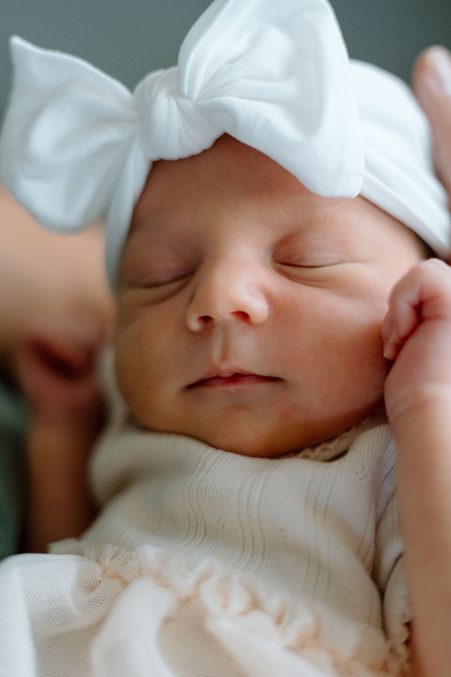 Sleeping newborn baby wearing a white bow headband and delicate white outfit.