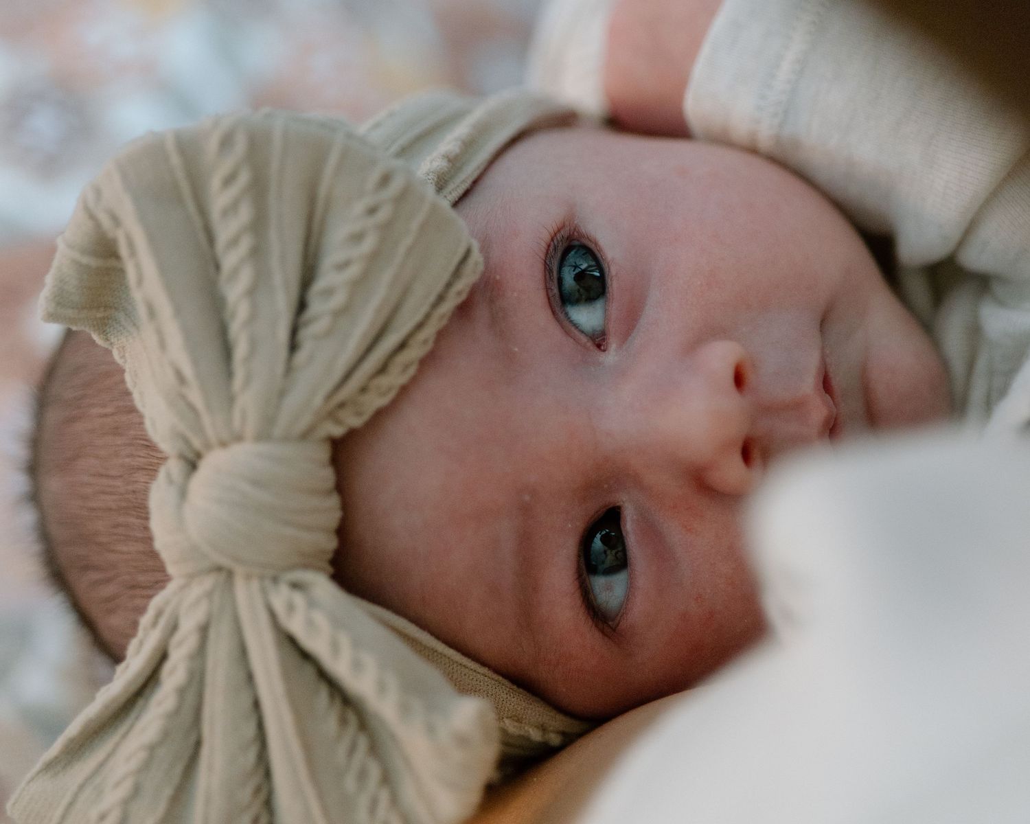 Baby with bright blue eyes wearing cream bow headband gazes sweetly at camera.