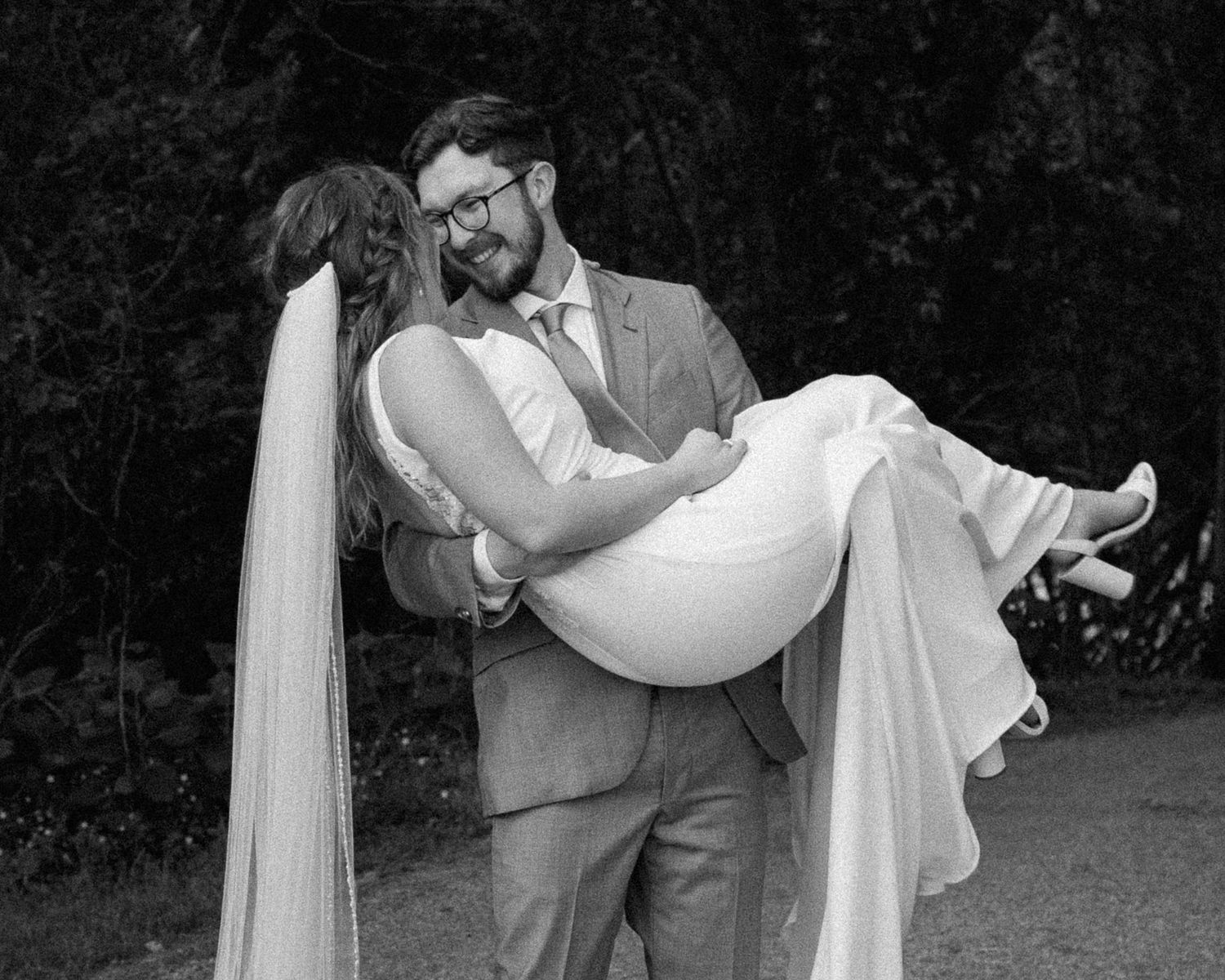 Romantic black and white portrait of newlyweds in a playful carrying pose outdoors.
