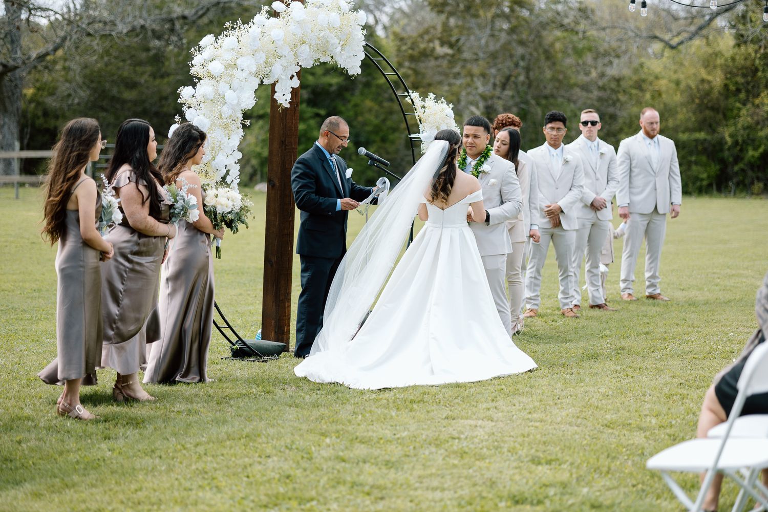 Garden wedding ceremony with guests in neutral colors gathered around a white floral arch.