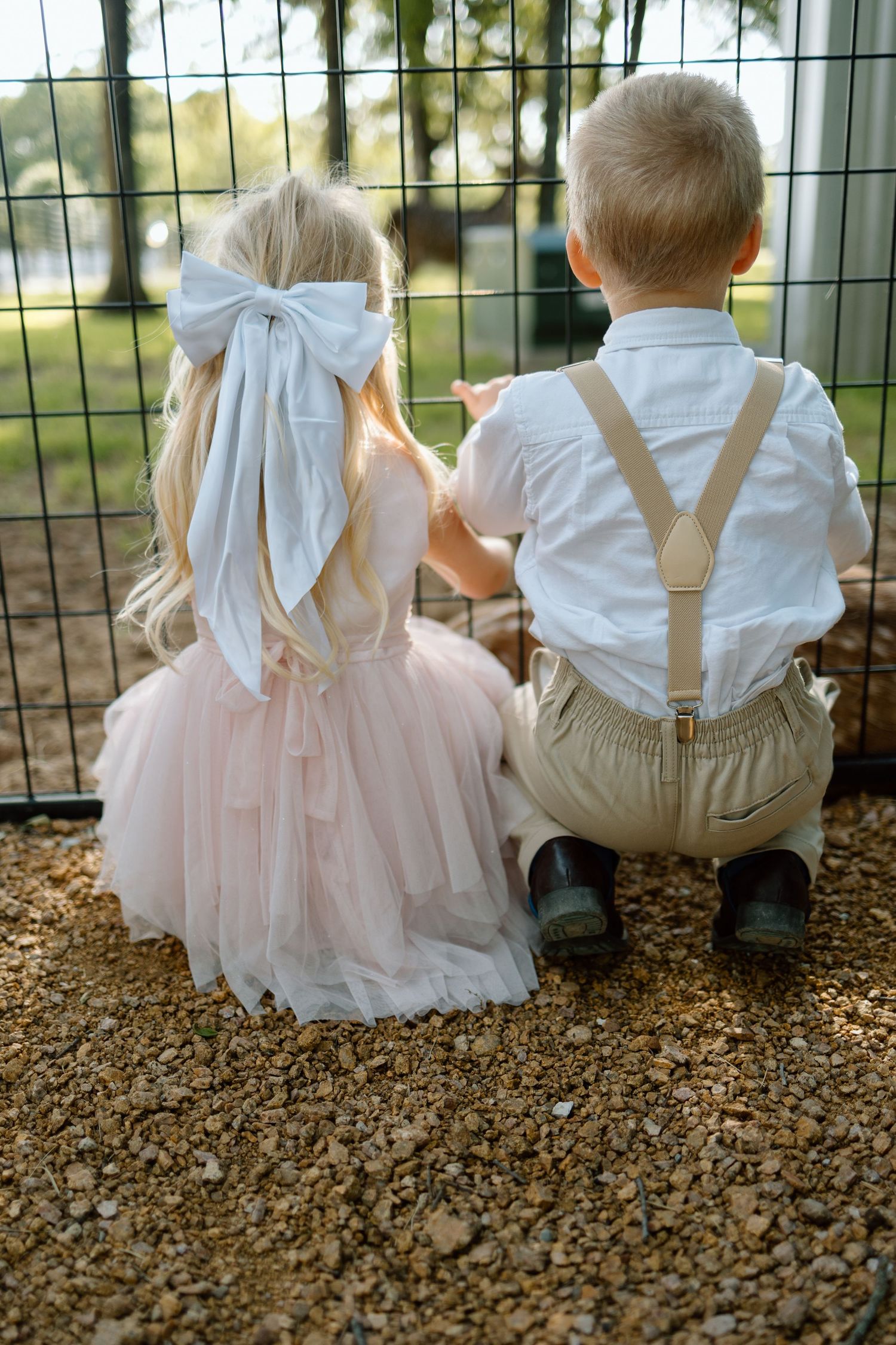 A flower girl in pink tulle and ring bearer in suspenders sit together looking through a fence.