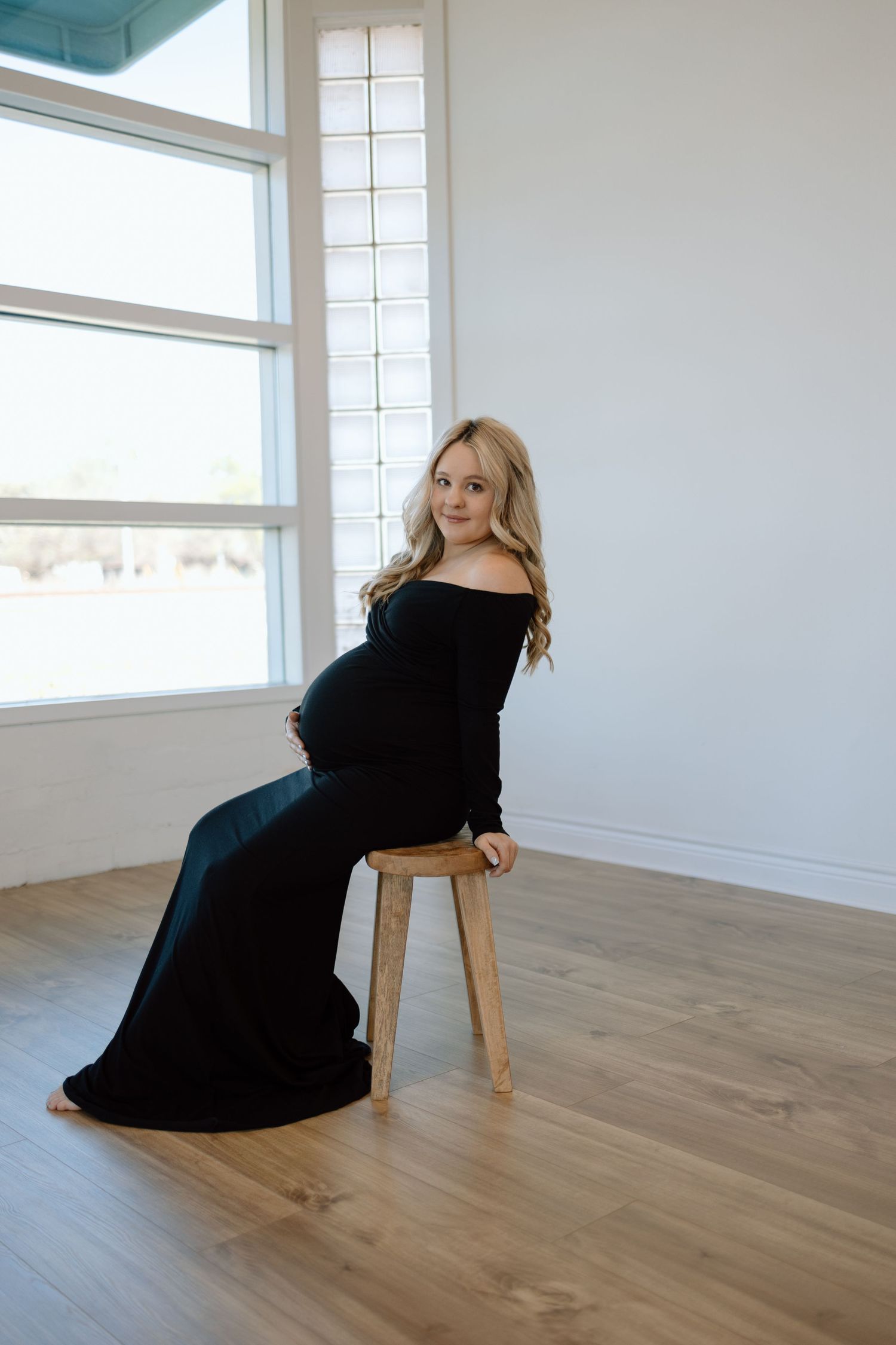 Maternity photo showing someone wearing a long black dress while posing on a wooden stool in a bright, minimalist room.