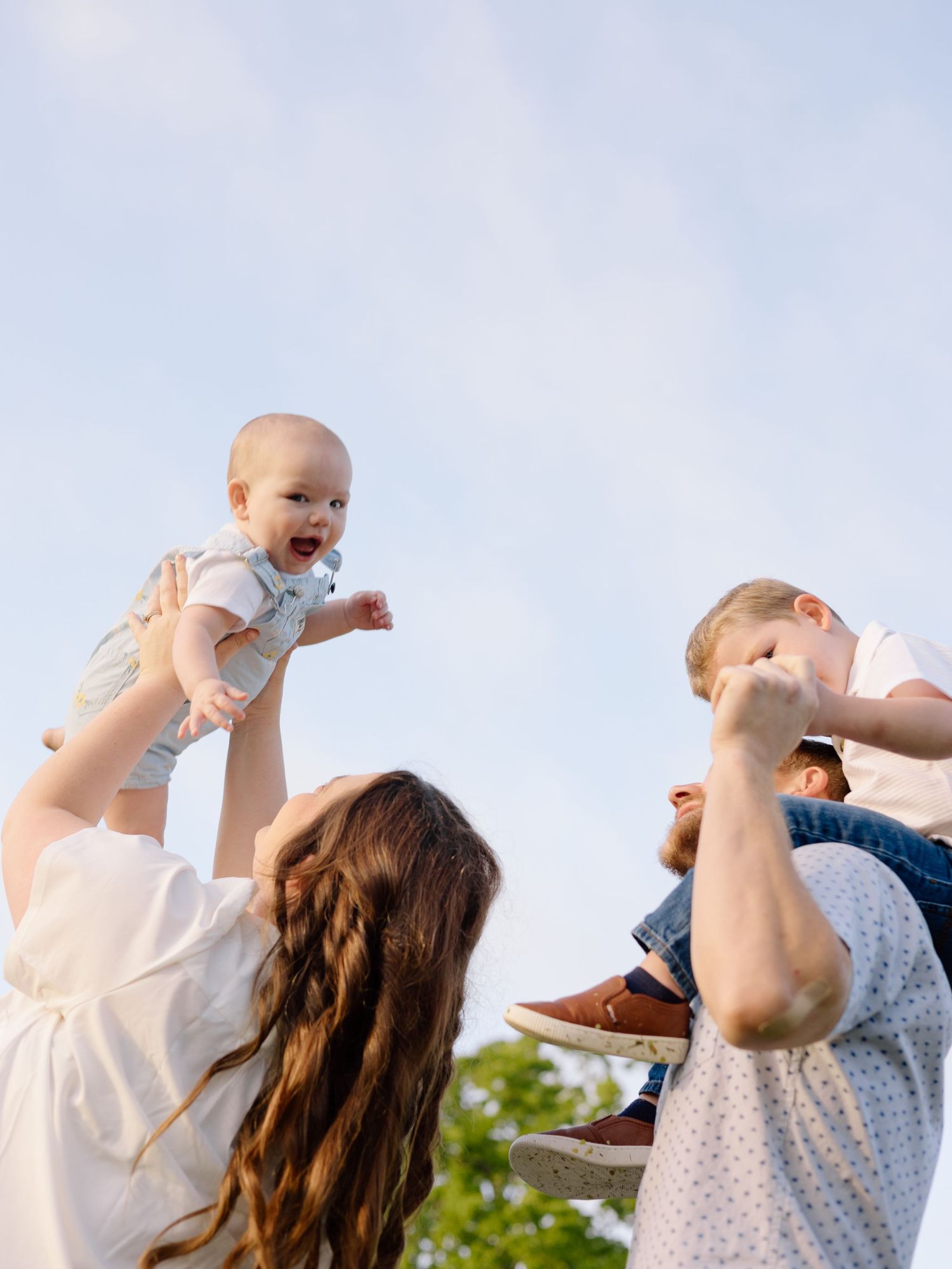 Two adults playfully lift a delighted baby into the sunny summer sky against a blue background with trees.