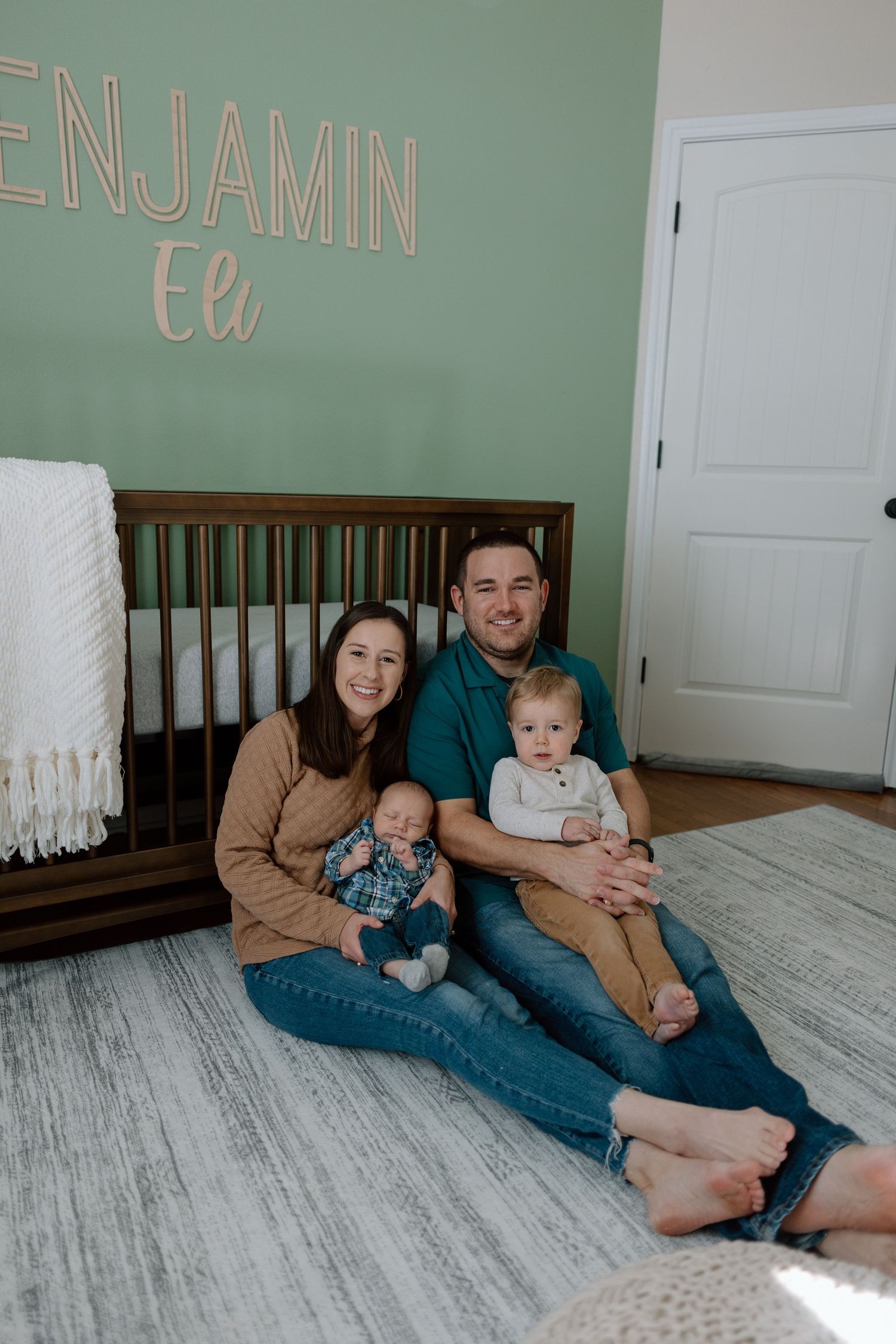 A nursery decorated with mint green walls and name letters features a wooden crib and family sitting on gray flooring.