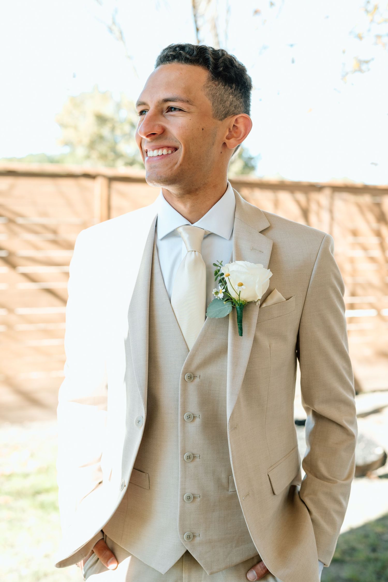 Groom in tan three-piece suit with white rose boutonniere smiling outdoors.