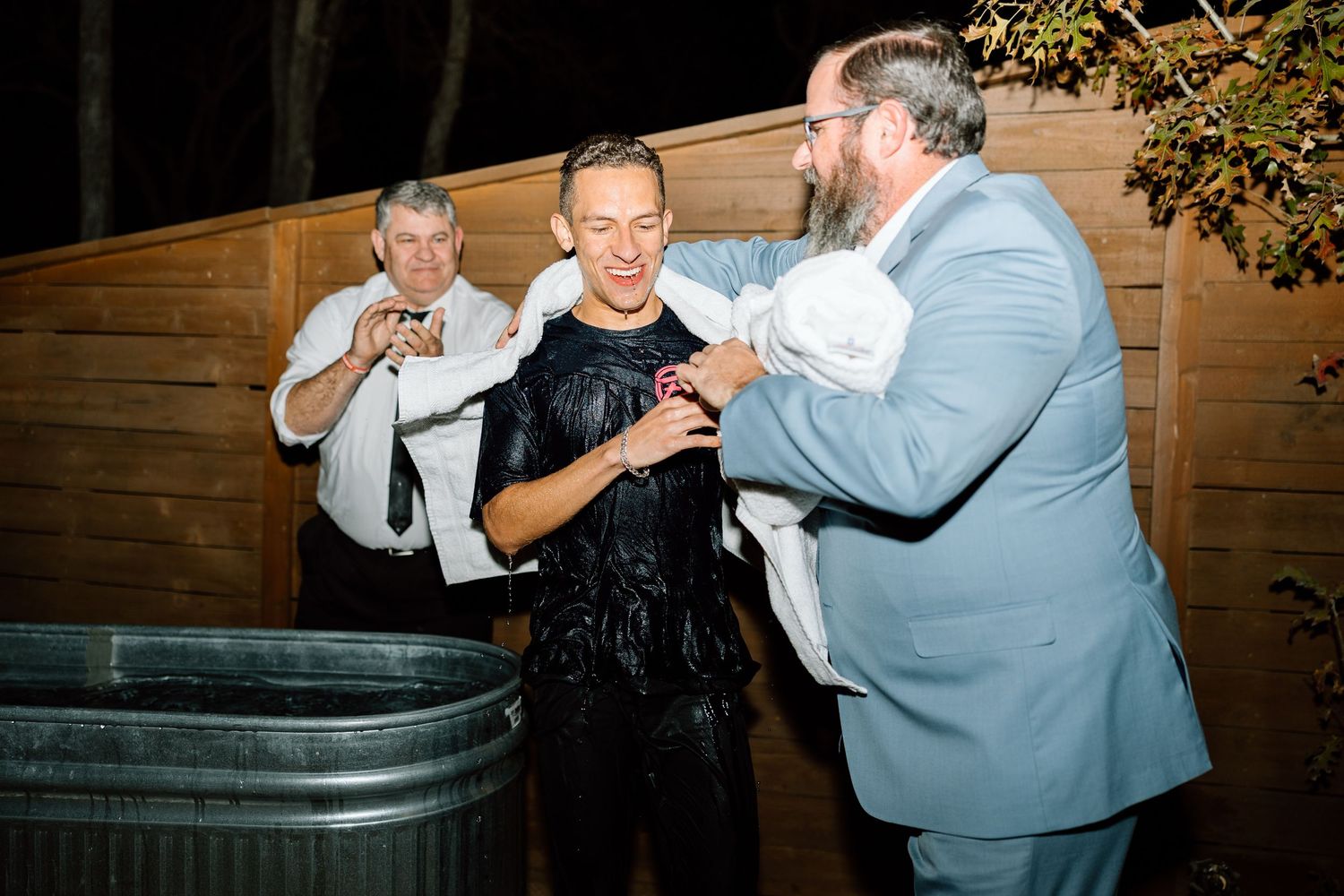 Man being helped out of metal trough after outdoor baptism at night.