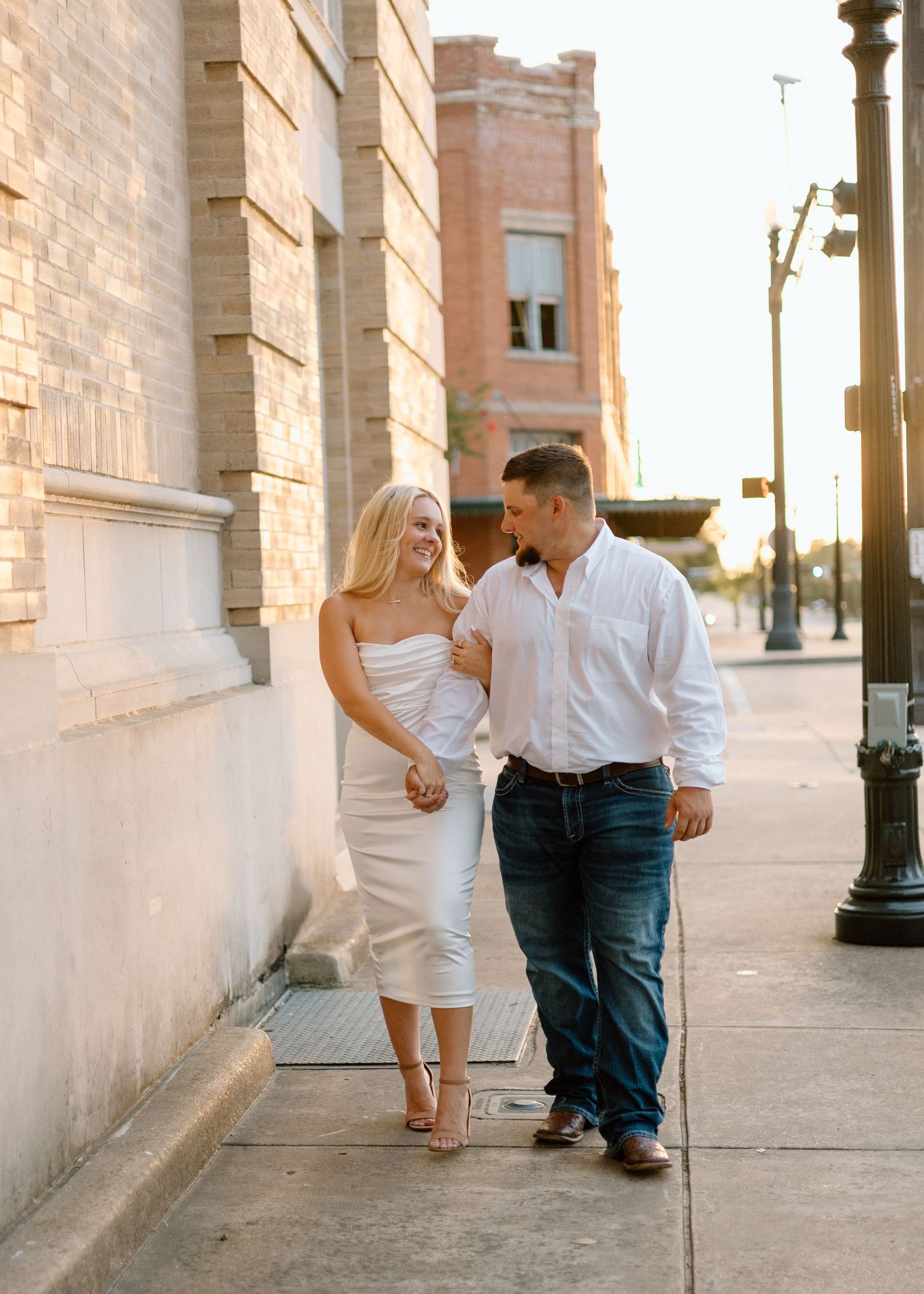 Couple walking hand-in-hand on a sunlit urban sidewalk, woman in white midi dress, man in jeans and white shirt.