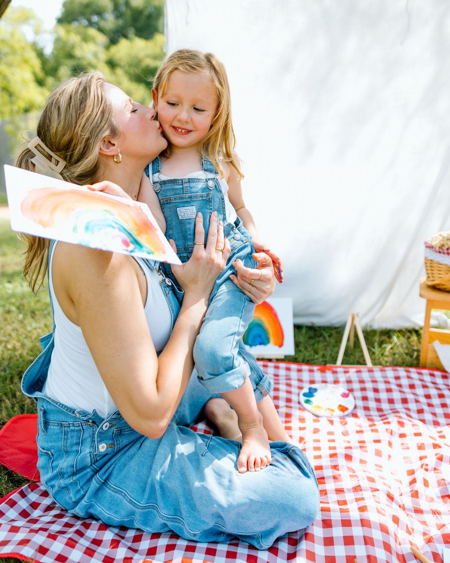 Mom kissing toddler girl in matching denim overalls on red gingham picnic blanket outdoors with rainbow artwork.