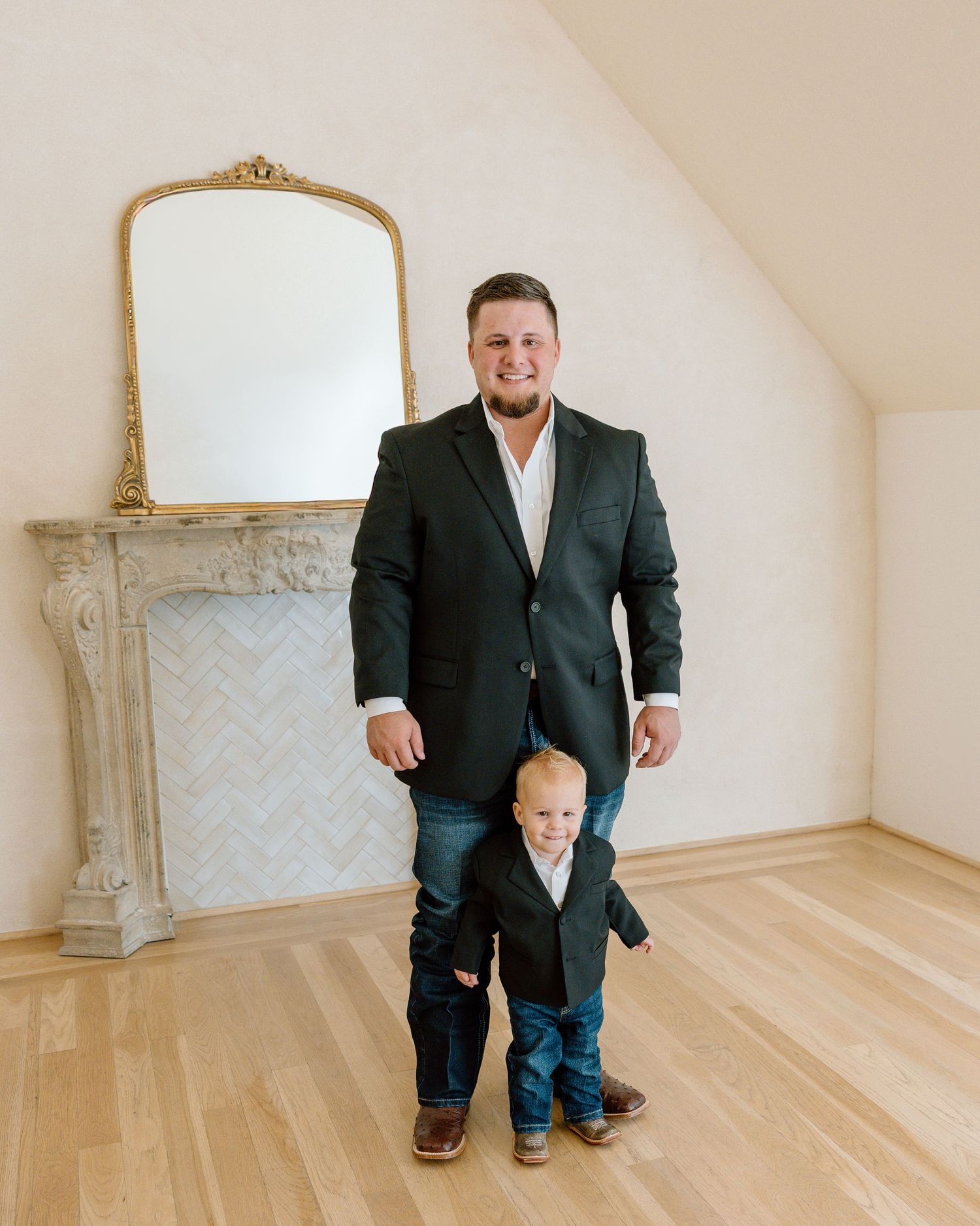 Man in black blazer and jeans smiling with a toddler boy in matching outfit before a decorative fireplace.
