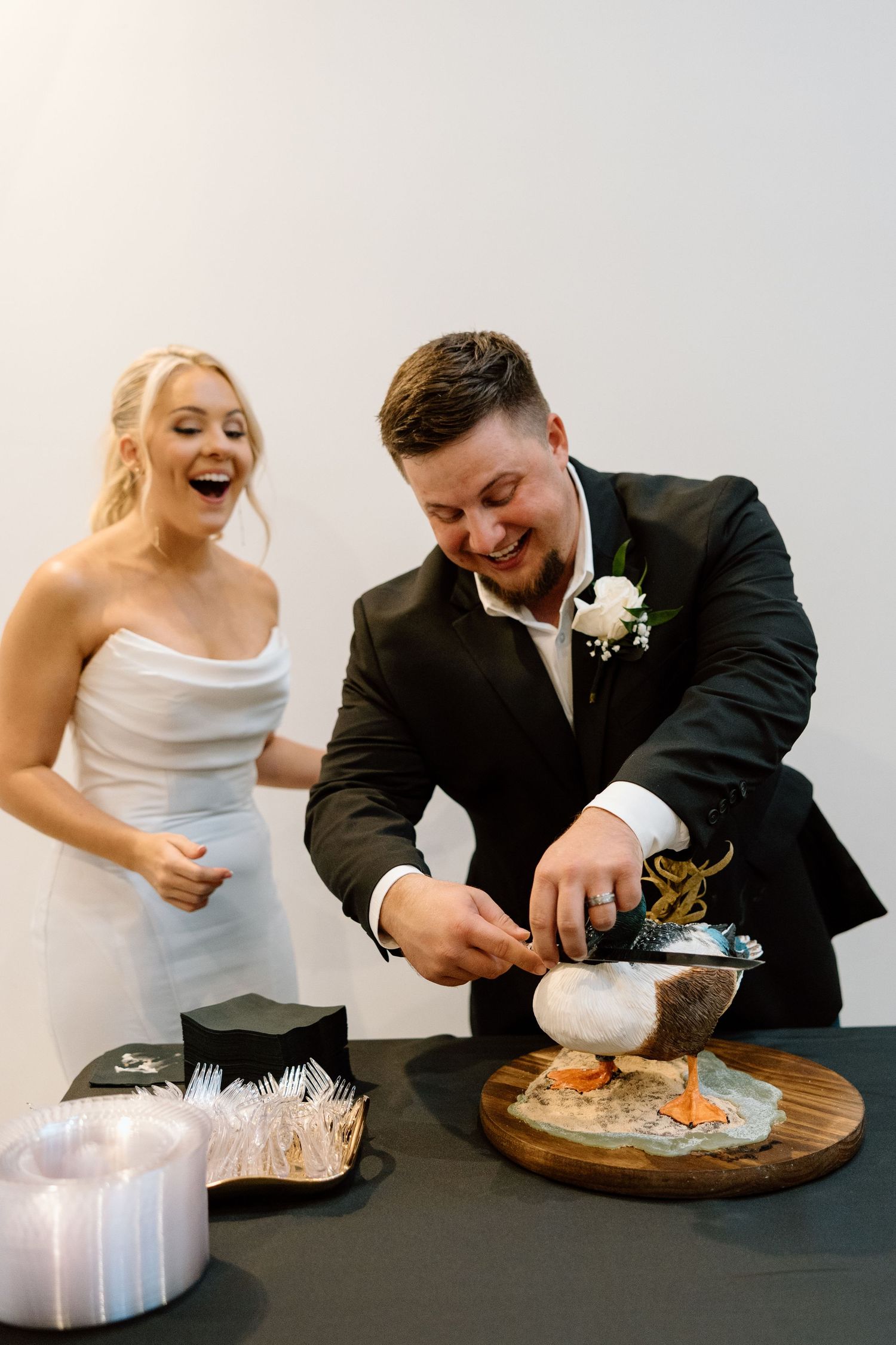 Bride and groom laughing while cutting a unique duck-shaped wedding cake on a wooden board.