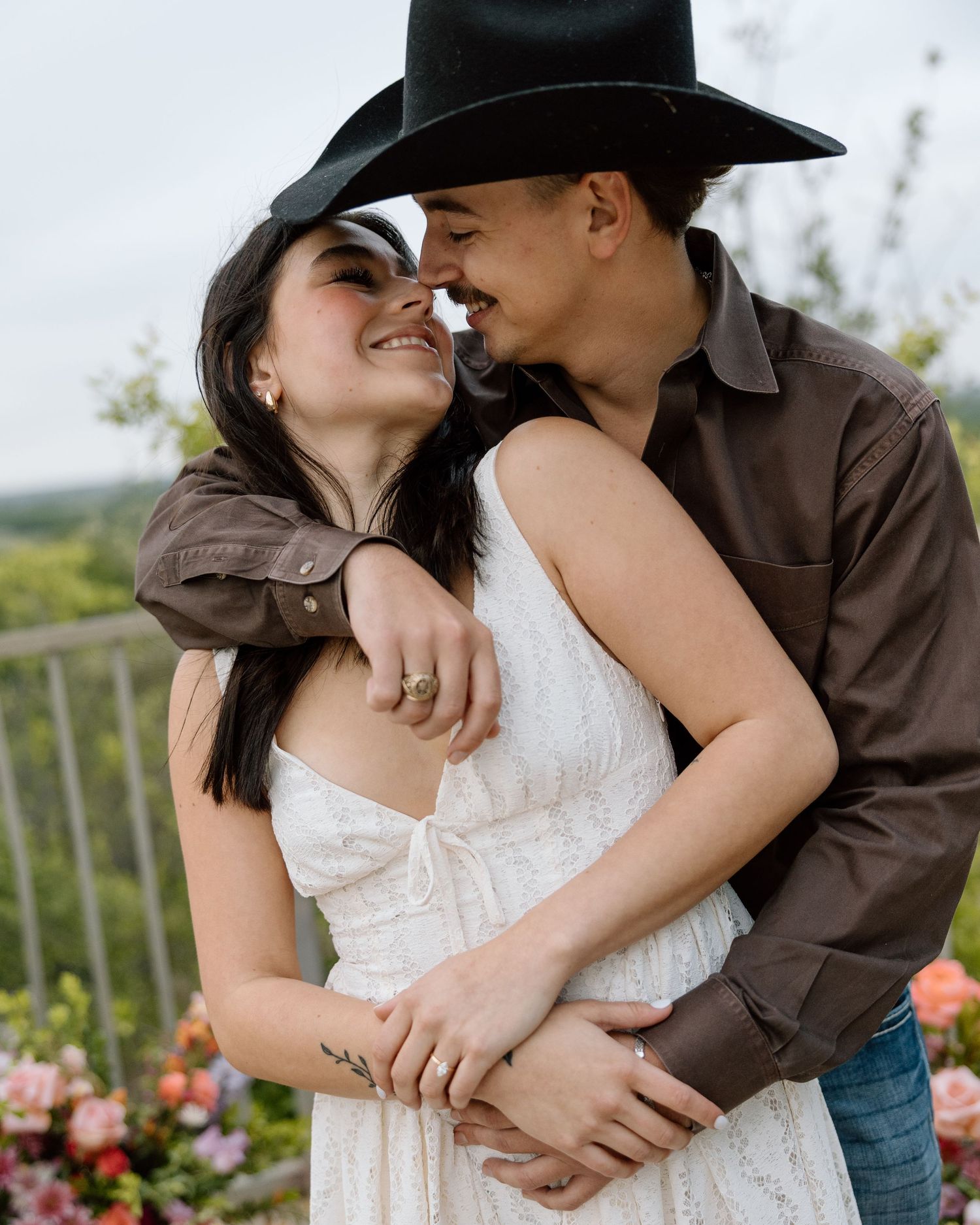 Couple in romantic embrace, woman in white dress and man in black cowboy hat, surrounded by flowers.