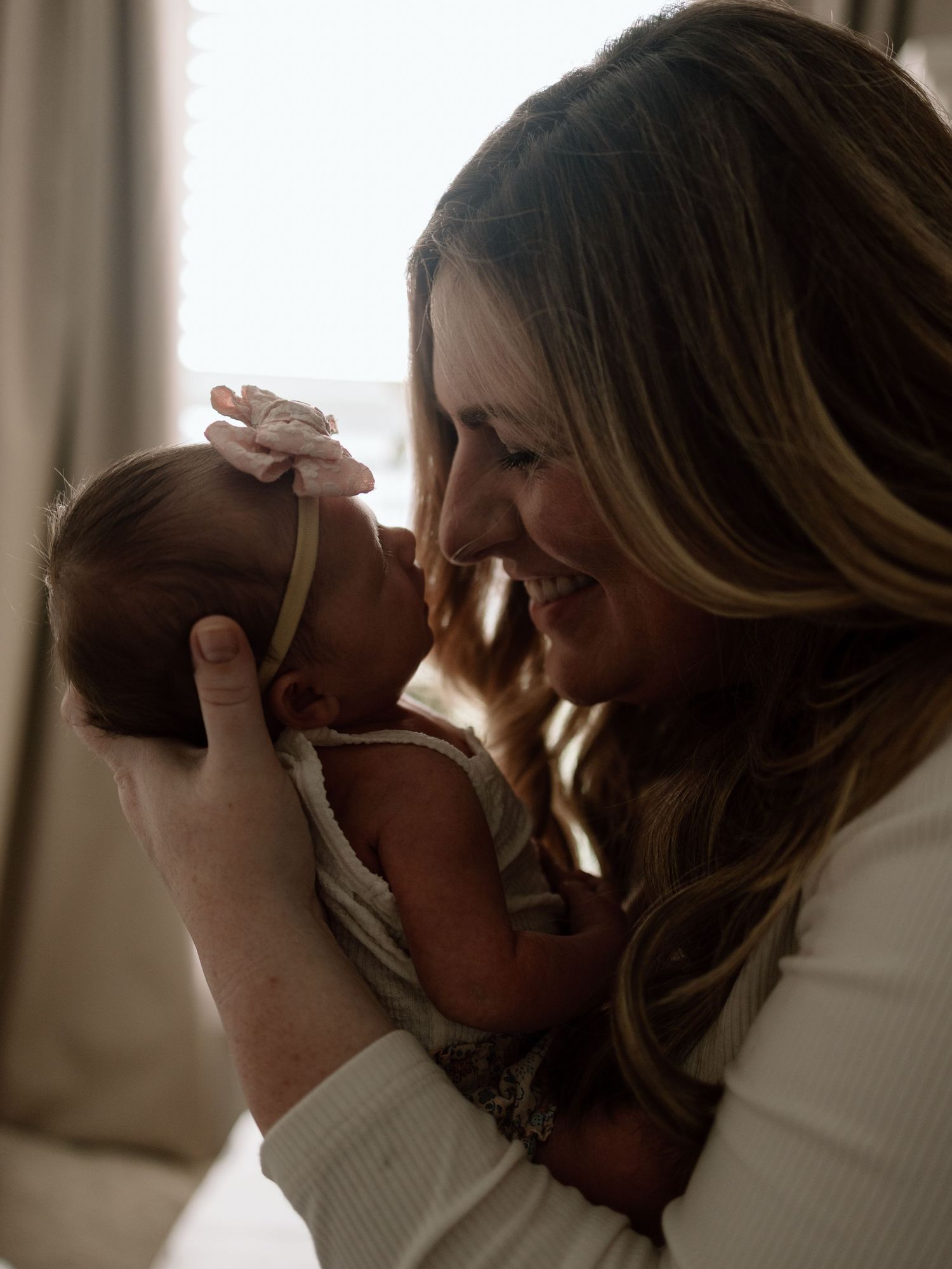 An intimate moment captured as a woman in white lovingly cradles and nuzzles a newborn baby in soft window light.