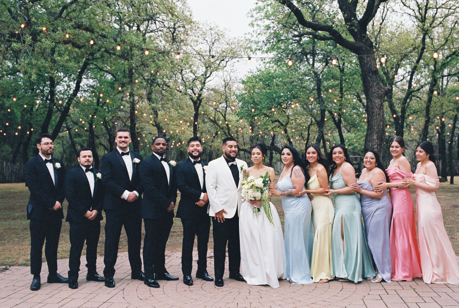 Large wedding party in formal attire poses together outdoors in wooded setting.