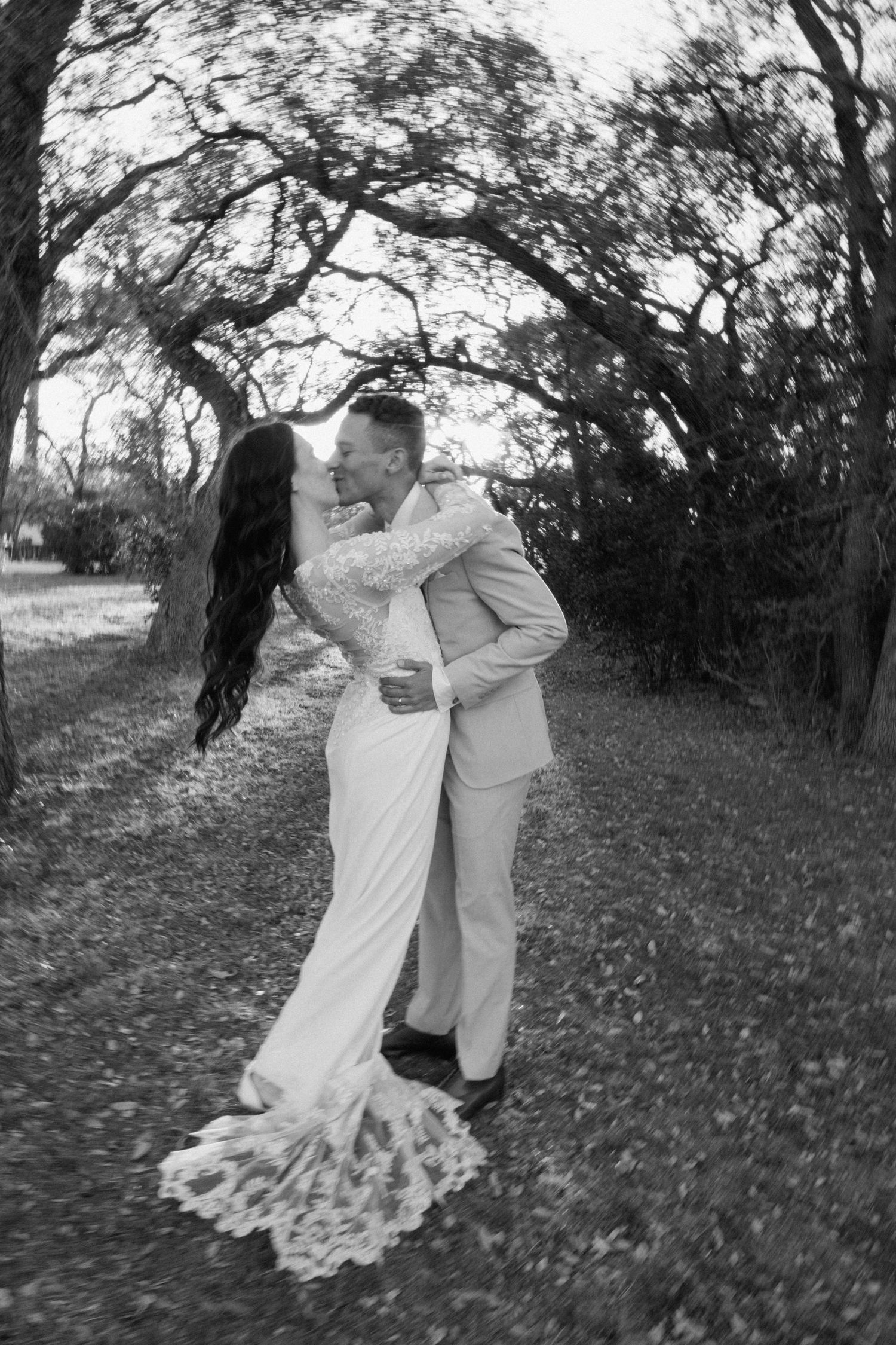 Black and white photo of a bride and groom sharing a kiss outdoors under trees on their wedding day.