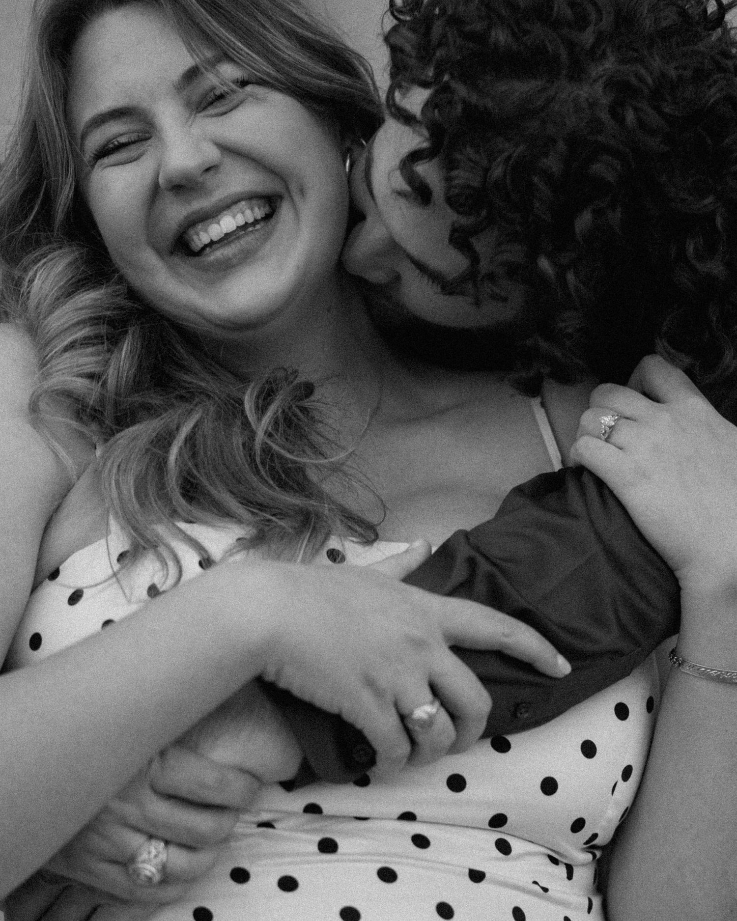 Black and white photo of two women laughing and embracing warmly, one wearing a polka dot dress.