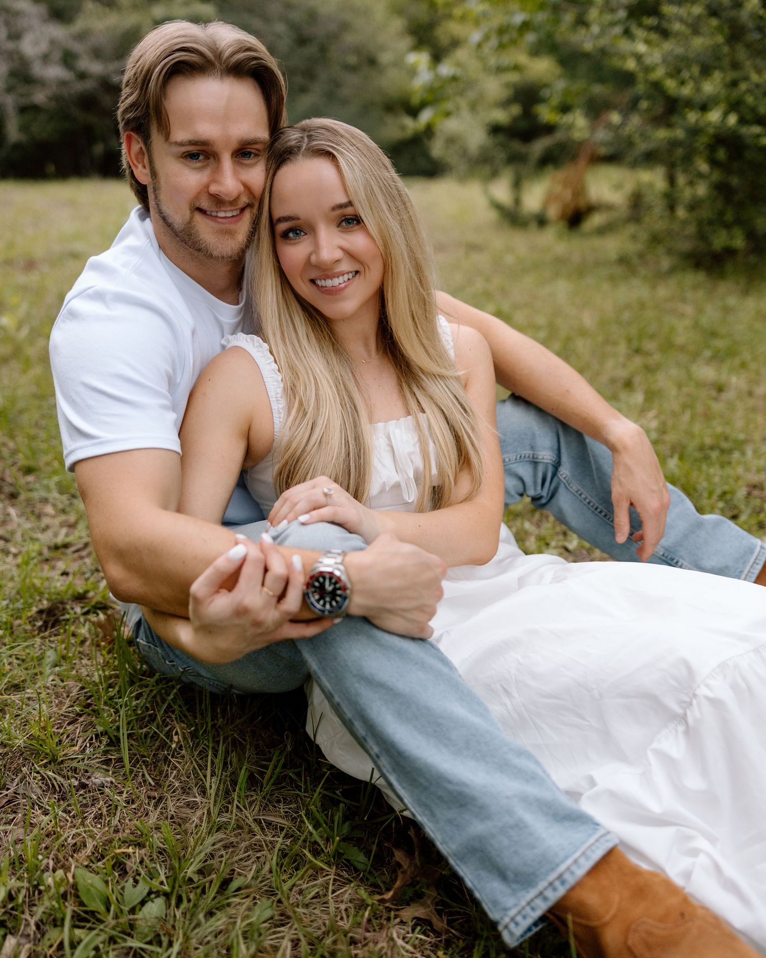 A smiling couple sits together on grass in a park, the woman in a white dress and the man in a white shirt.