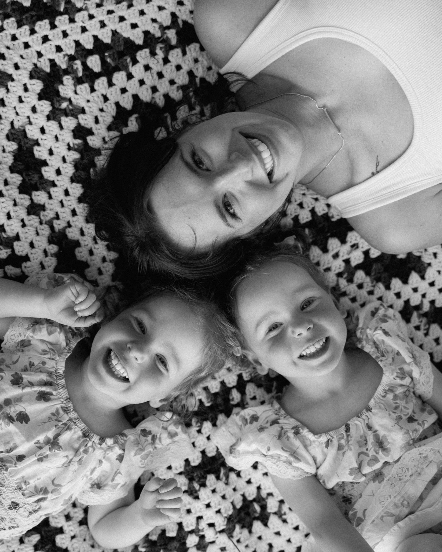Black and white photo of a smiling mother lying on a crochet blanket with her two young daughters.