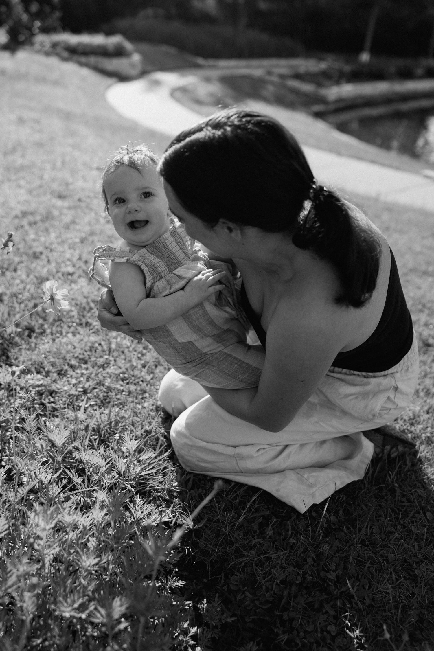 Black and white photo of a mother crouching on grass, lovingly holding her smiling baby outdoors near a garden path.