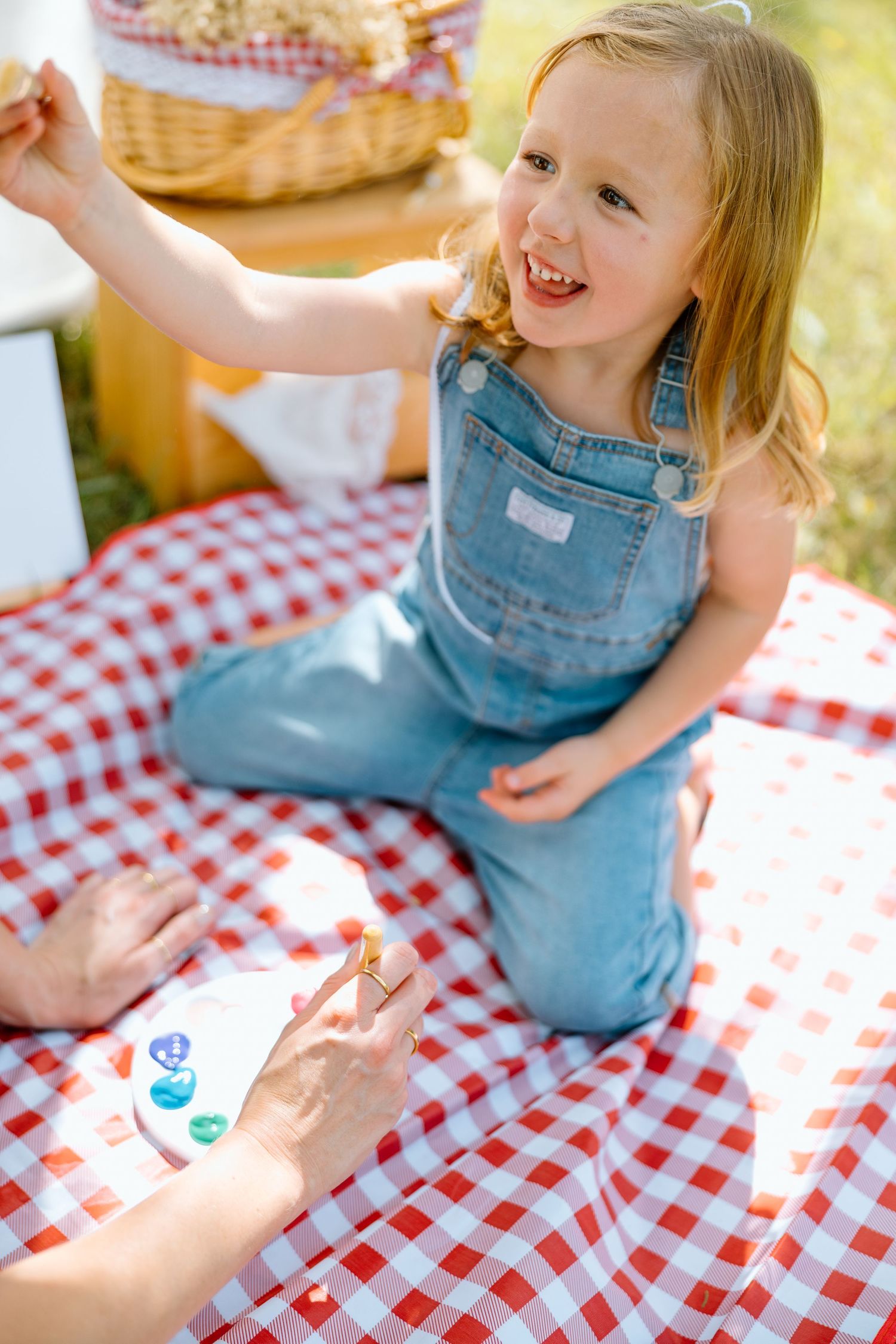 Happy little girl in denim overalls sitting on red checkered blanket at outdoor picnic, reaching up joyfully.