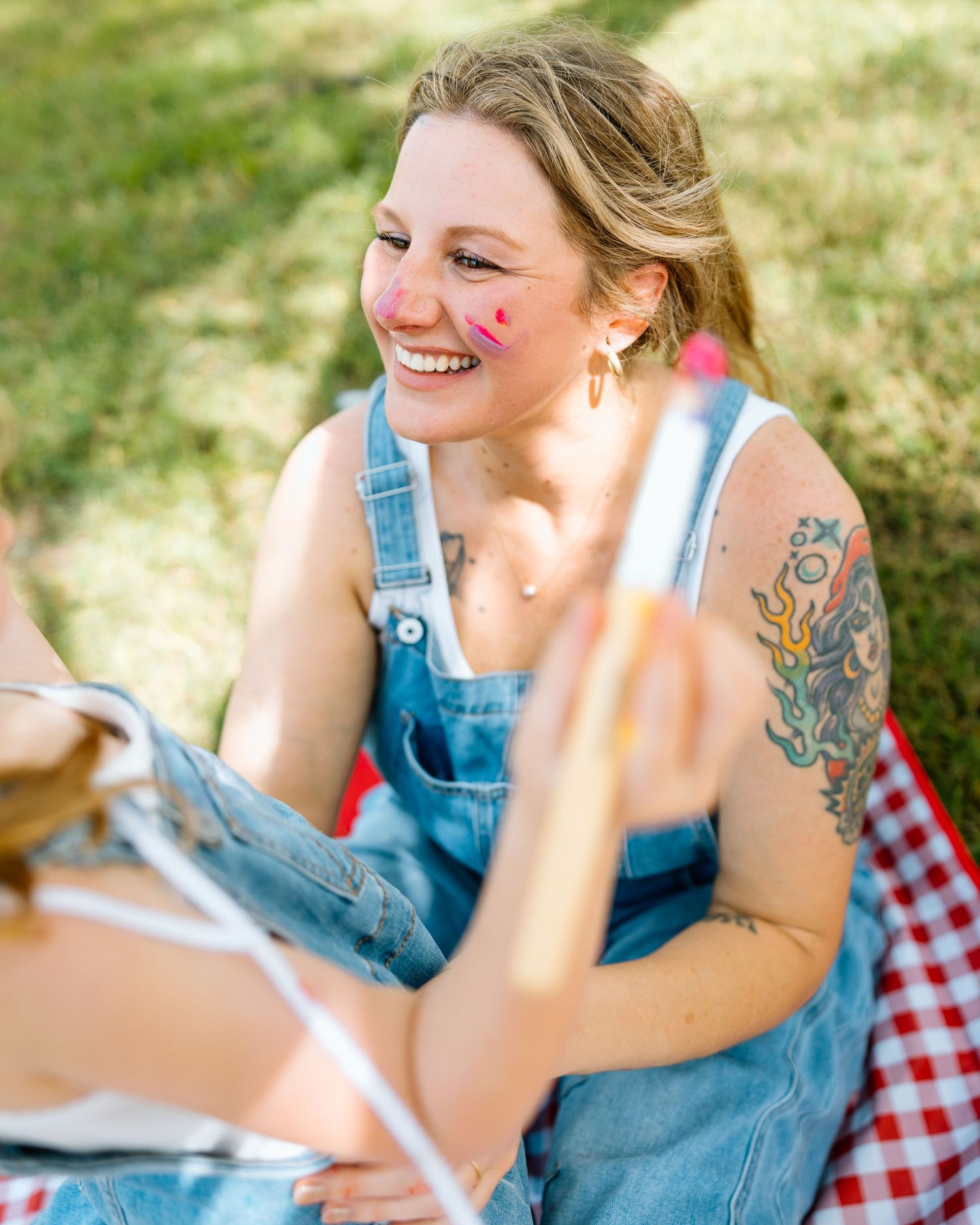 Smiling woman in denim overalls with face paint and tattoo enjoys outdoor picnic on red checkered blanket.