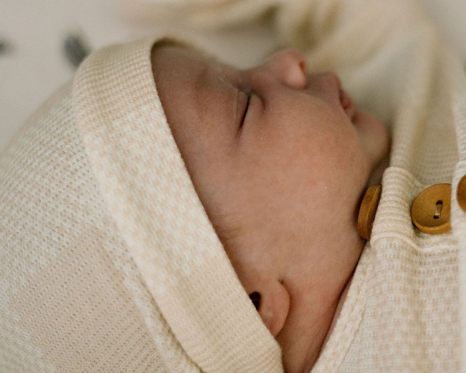 Peaceful newborn baby sleeping wrapped in a cream knit swaddle blanket with golden buttons.