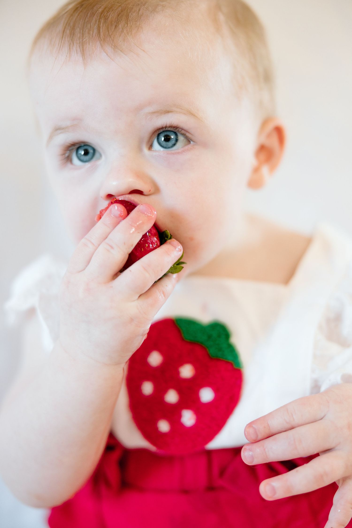 Blue-eyed baby girl in red strawberry outfit eating a fresh strawberry.