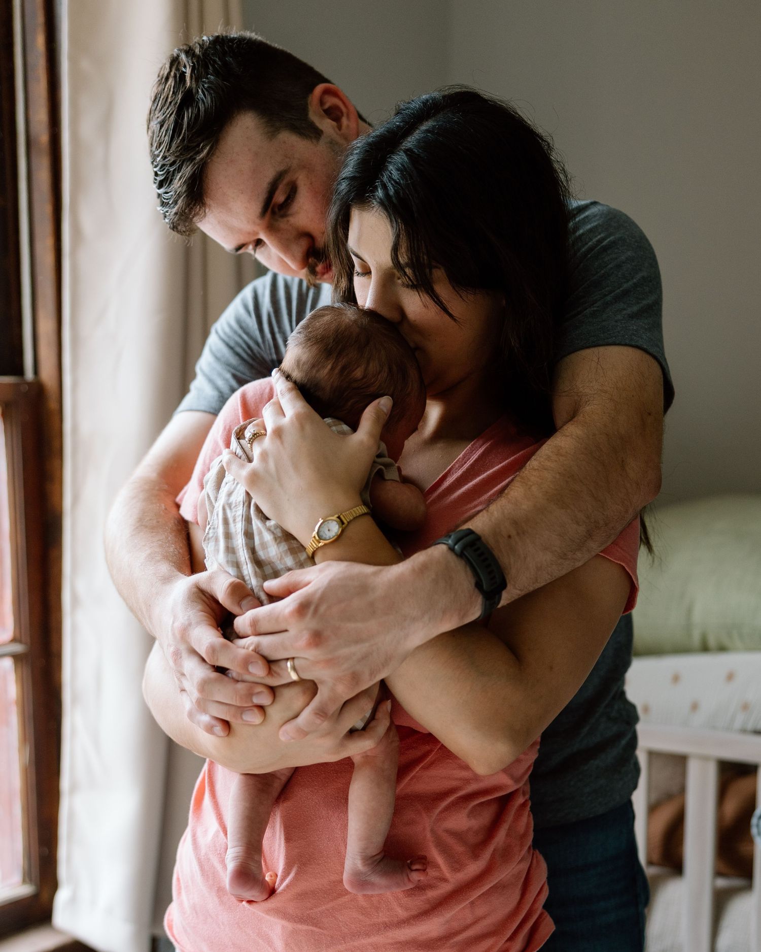 New parents tenderly holding and kissing their newborn baby by a bright window at home.