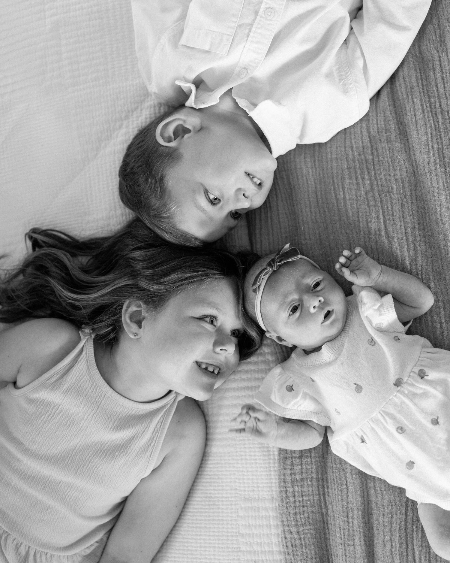 Black and white photo of three siblings lying together, two older girls smiling and a baby looking up.