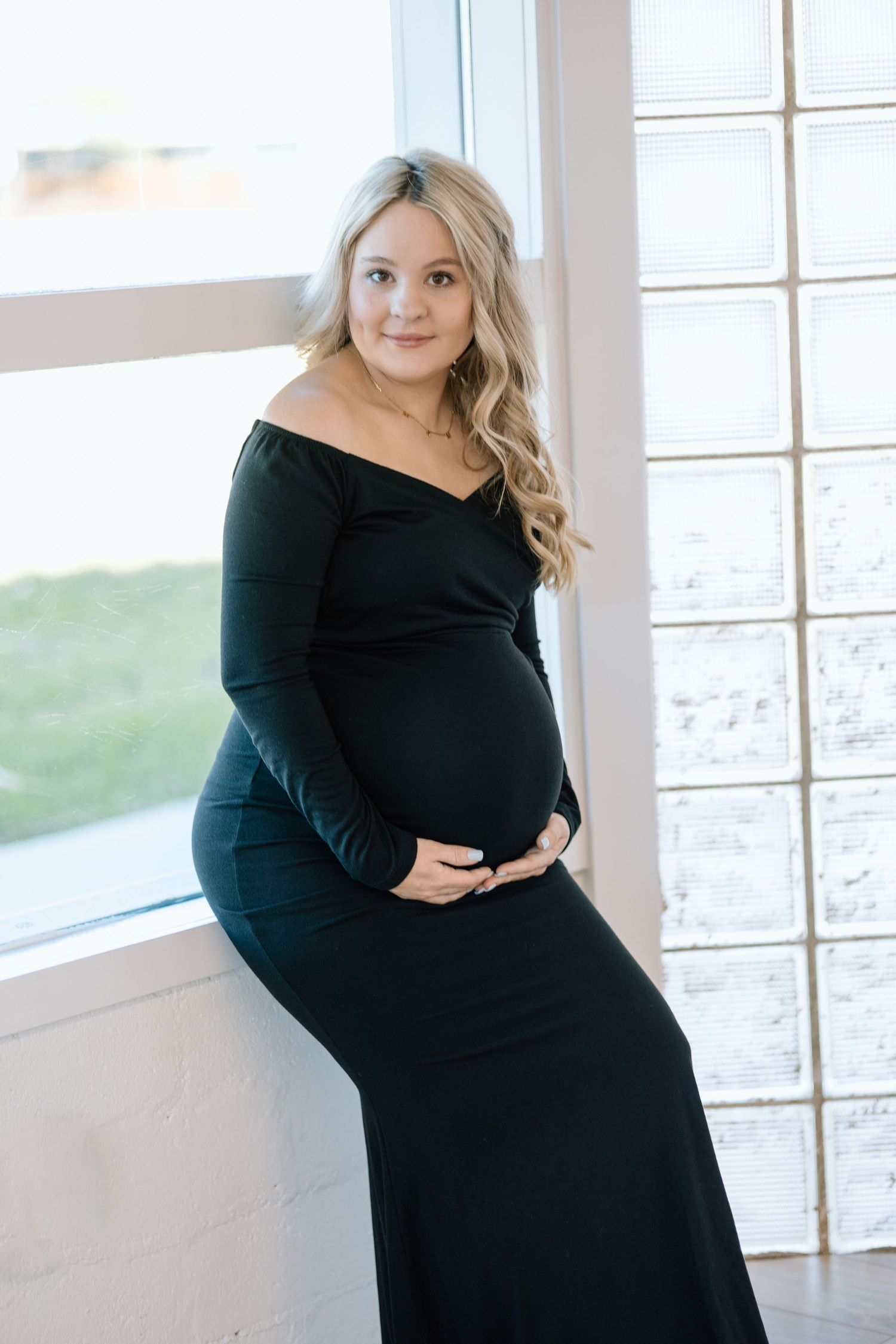 An expectant mother in a black off-shoulder maternity gown poses elegantly by a bright window in a natural light setting.