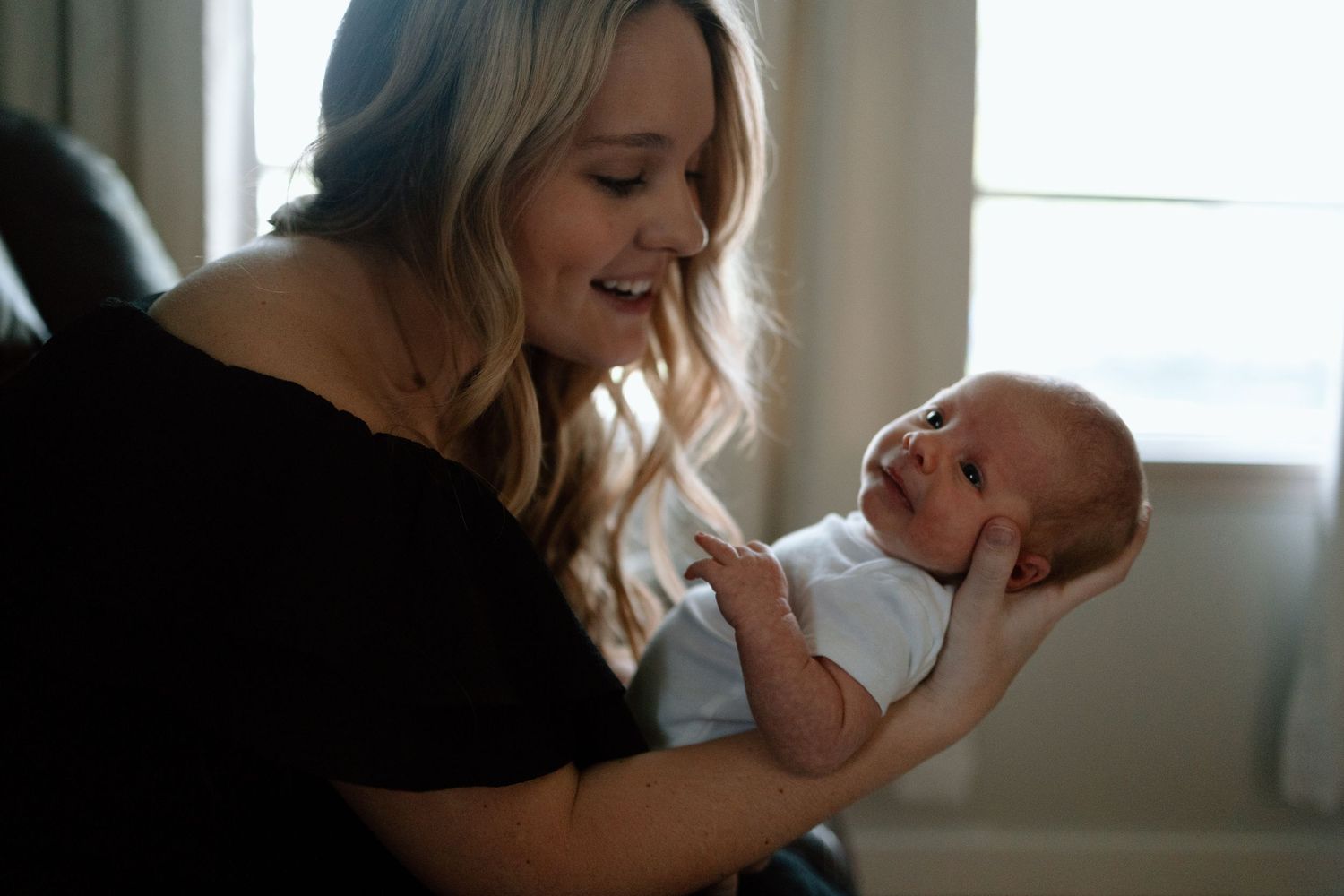 A parent in black clothing holds and interacts lovingly with a newborn baby in a bright sunlit room.