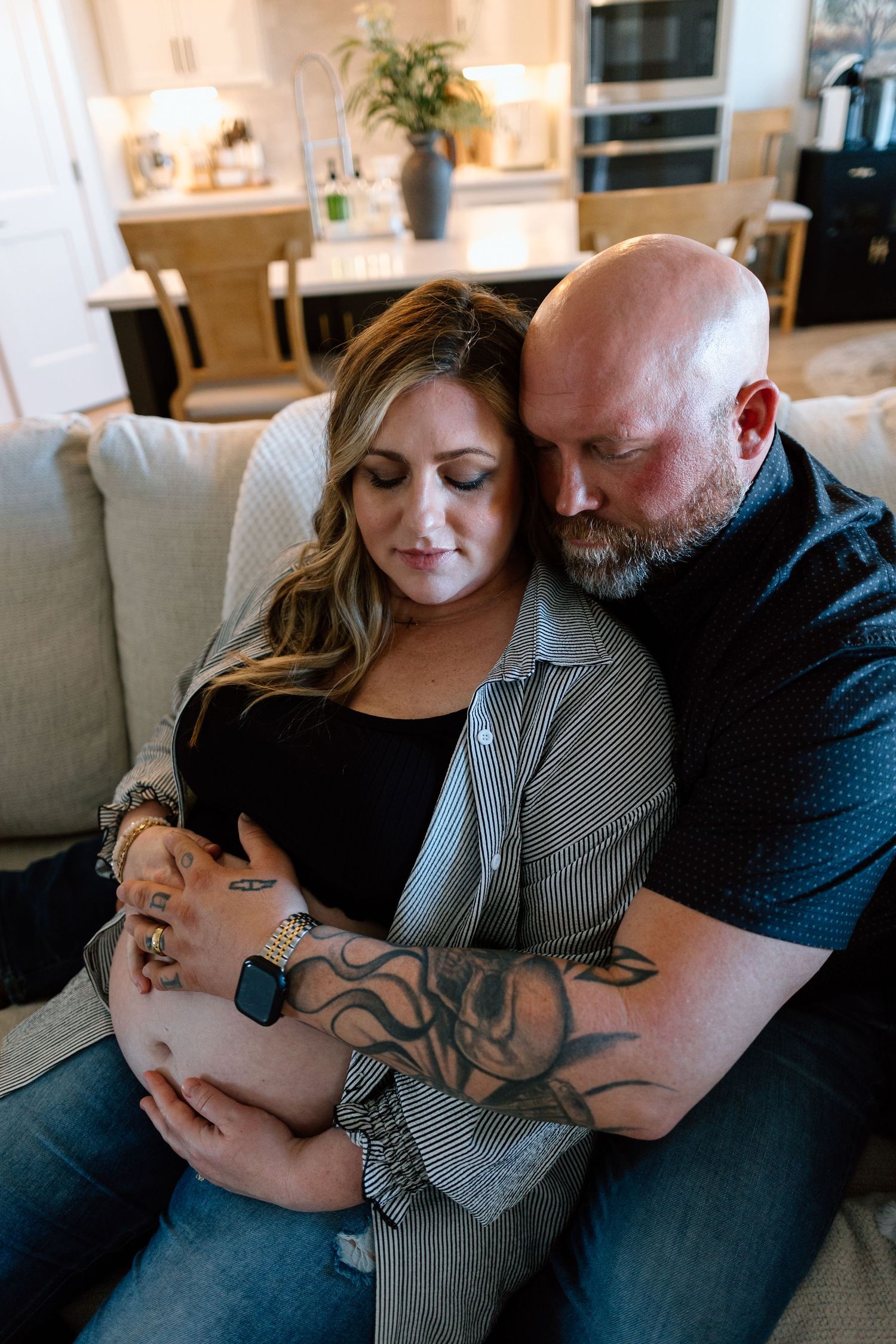 A couple shares an intimate moment on a couch, with tattooed arms wrapped in embrace.