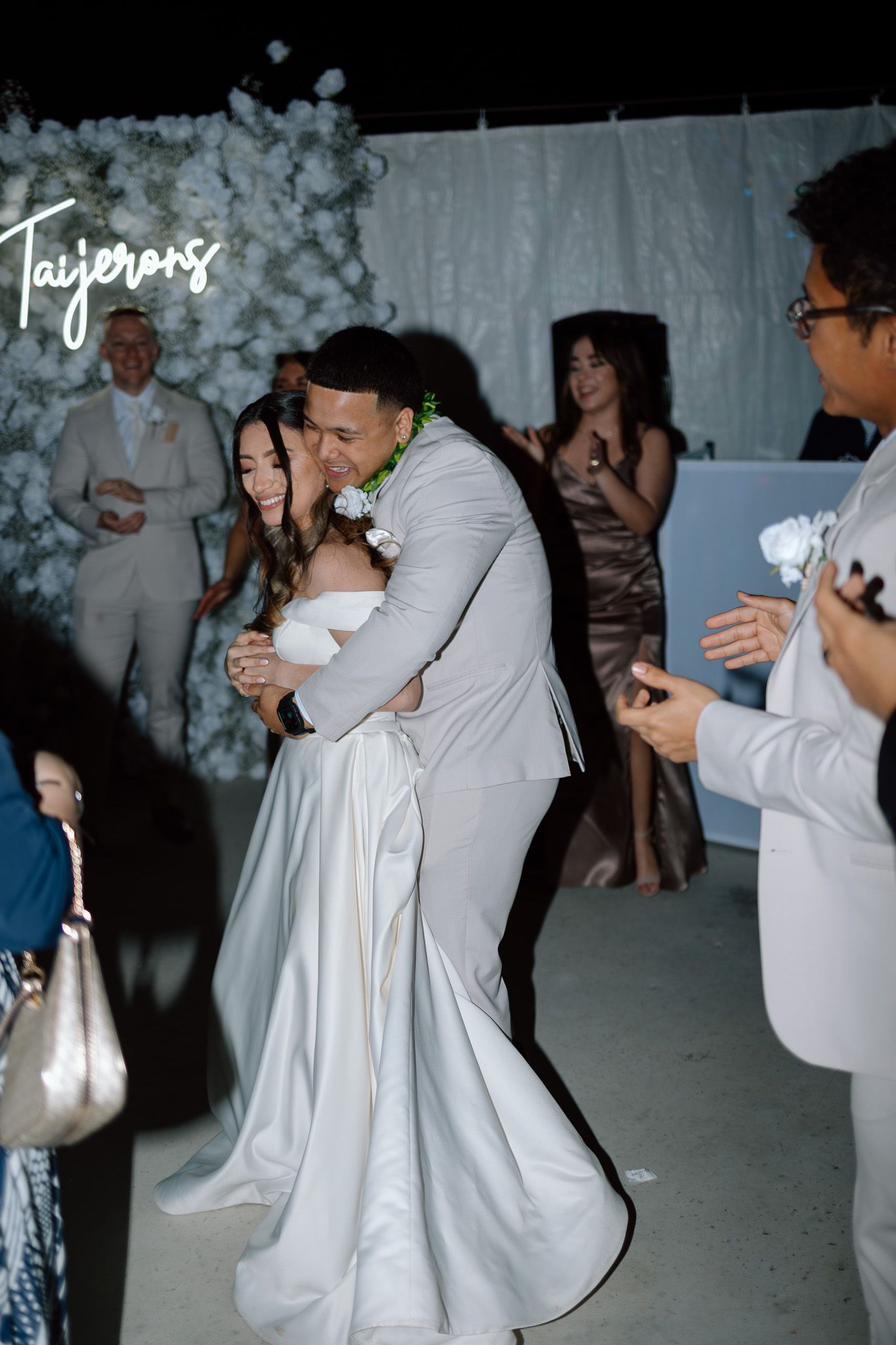 Wedding couple embracing on dance floor with white floral backdrop and neon signage.