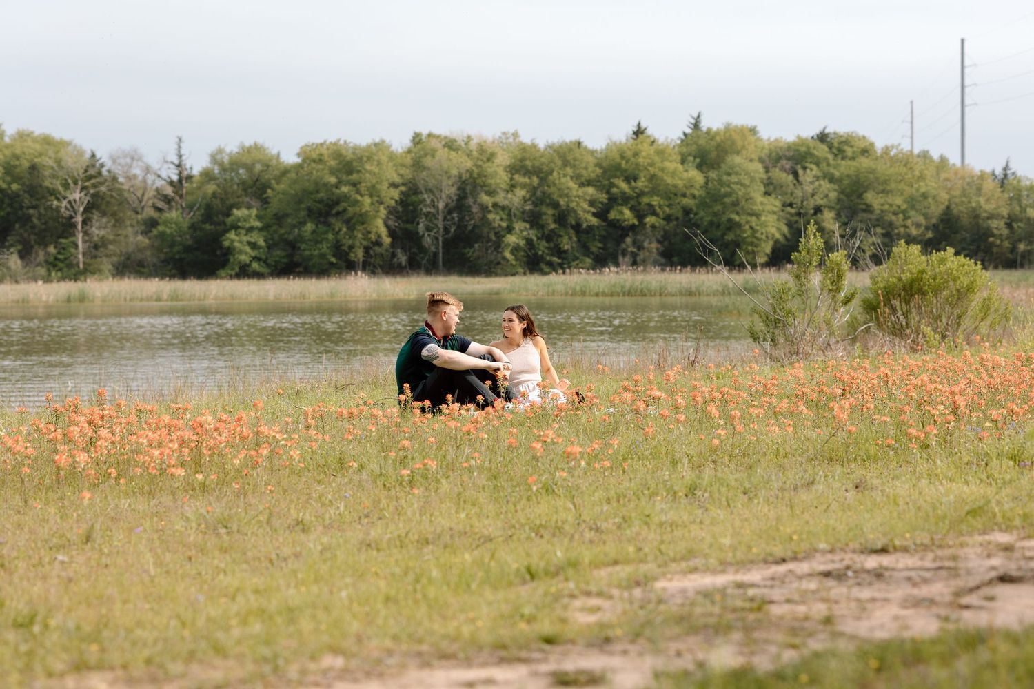 A couple enjoys a picnic together in a serene lakeside field dotted with orange flowers.