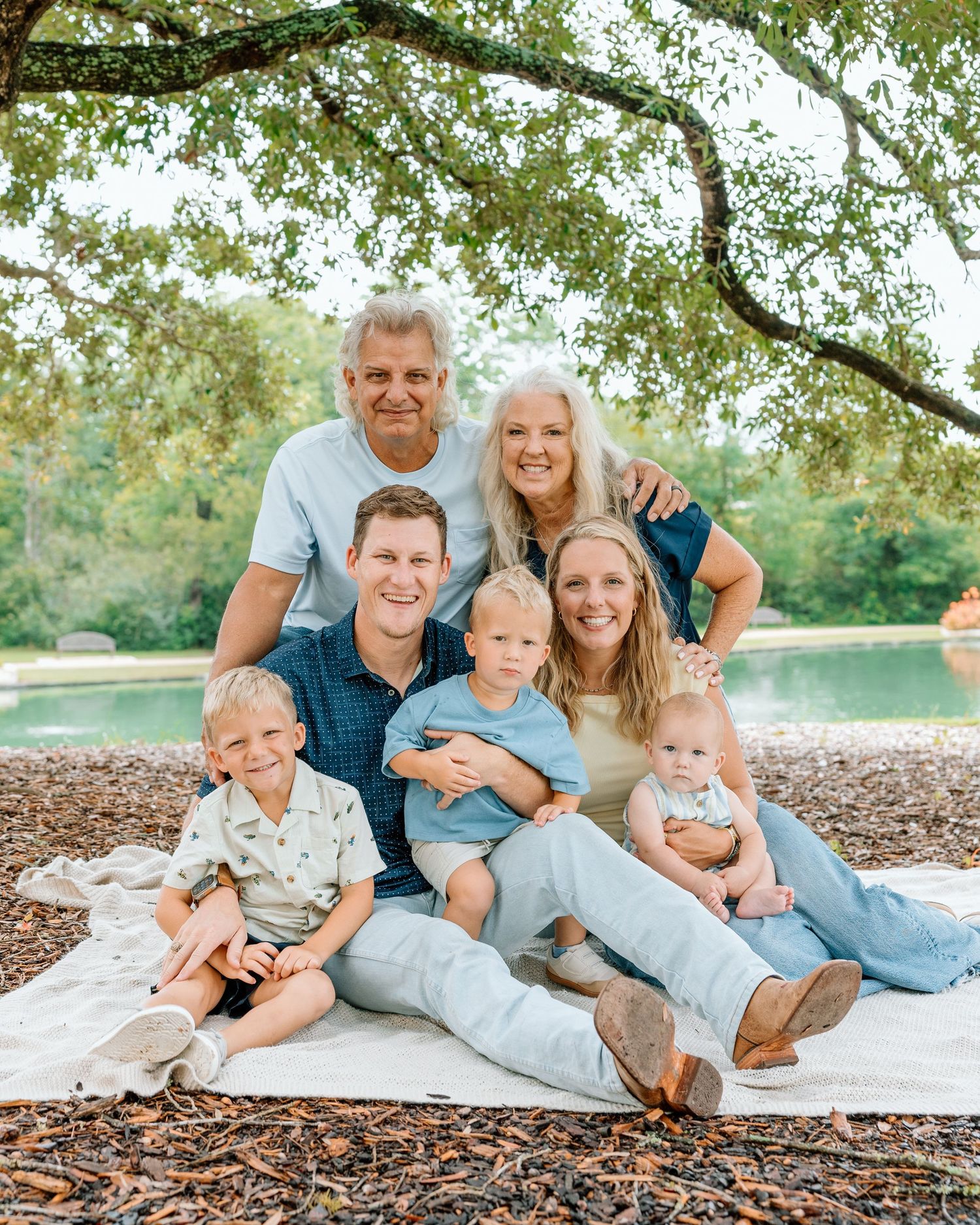 A multigenerational family of seven smiles together on a blanket outdoors near a pond under a large tree.