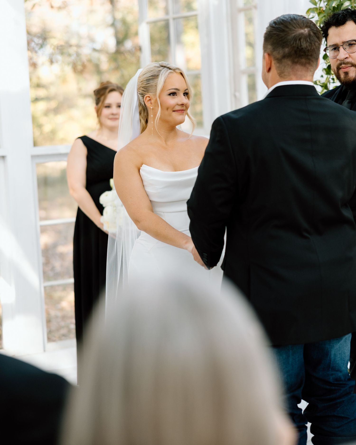 Smiling bride in strapless white gown exchanges vows with groom at intimate greenhouse wedding ceremony.