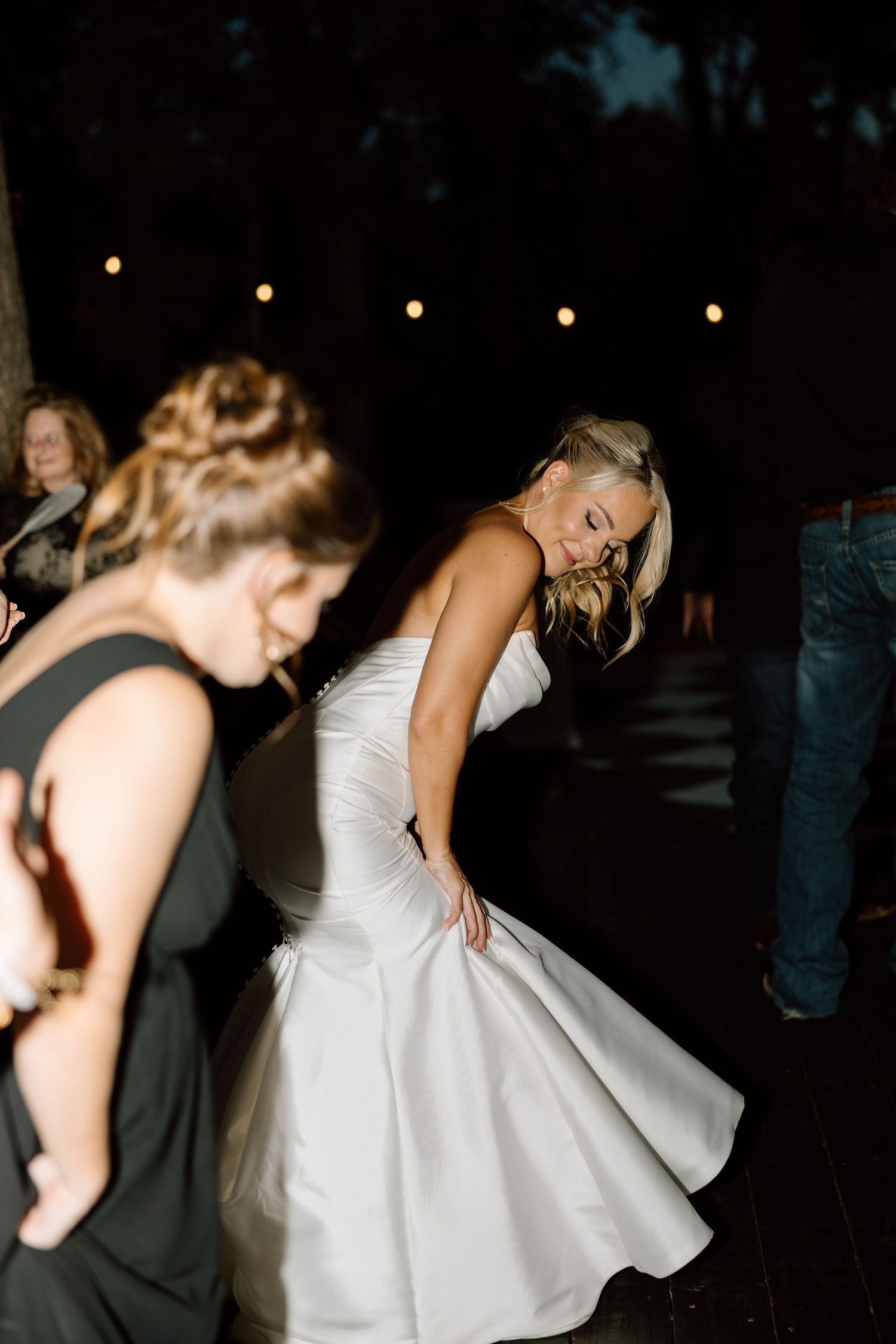 Bride in strapless white mermaid gown dances energetically under string lights at nighttime wedding reception.