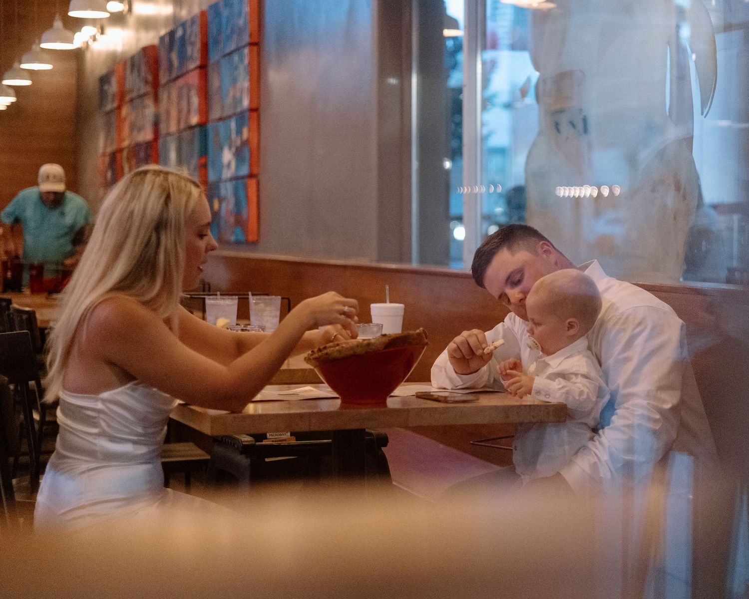 Newlywed couple in white wedding attire dining at a restaurant booth with their baby, sharing a casual post-wedding meal.