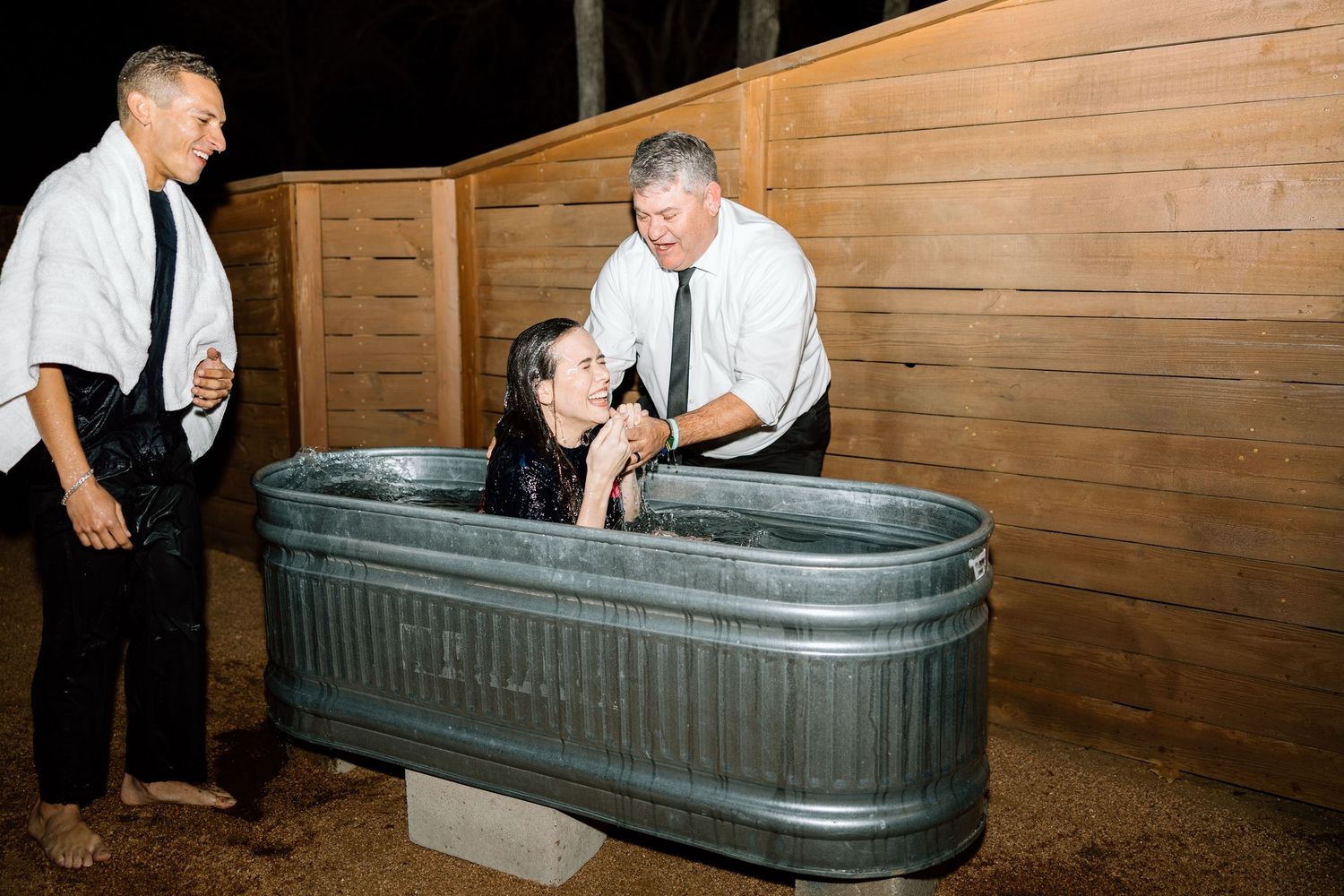 Woman being baptized in metal trough outdoors at nighttime ceremony.
