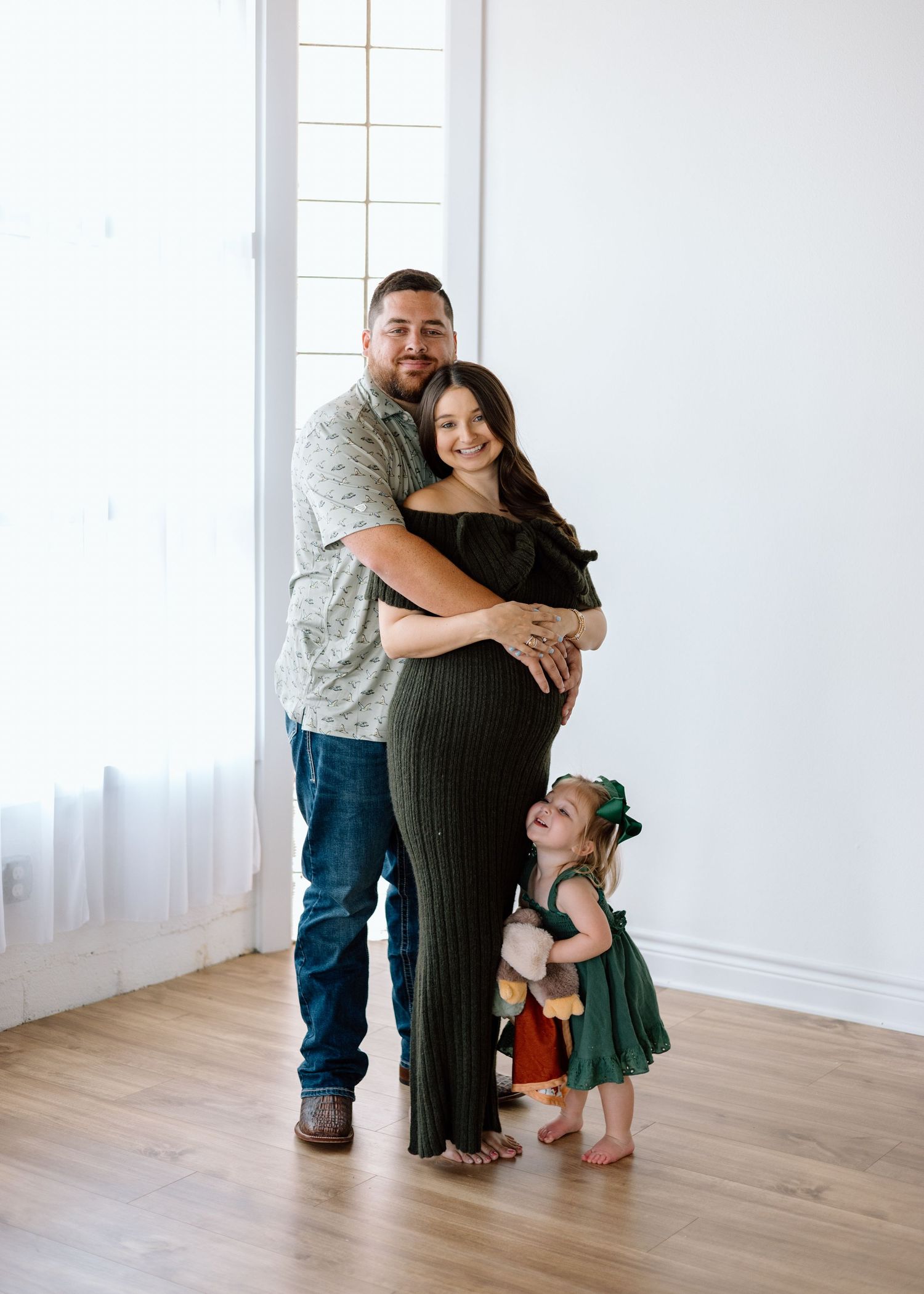 Pregnant woman in green dress embraced by man, with toddler girl in green dress standing beside them in bright studio.