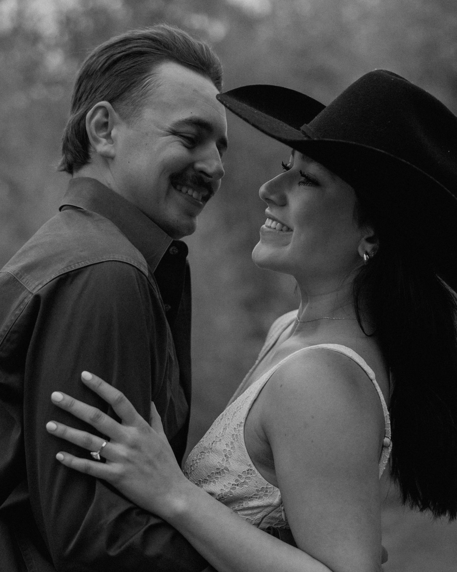 Black and white photo of a smiling couple embracing outdoors, woman wearing a cowboy hat and man in a dark shirt.