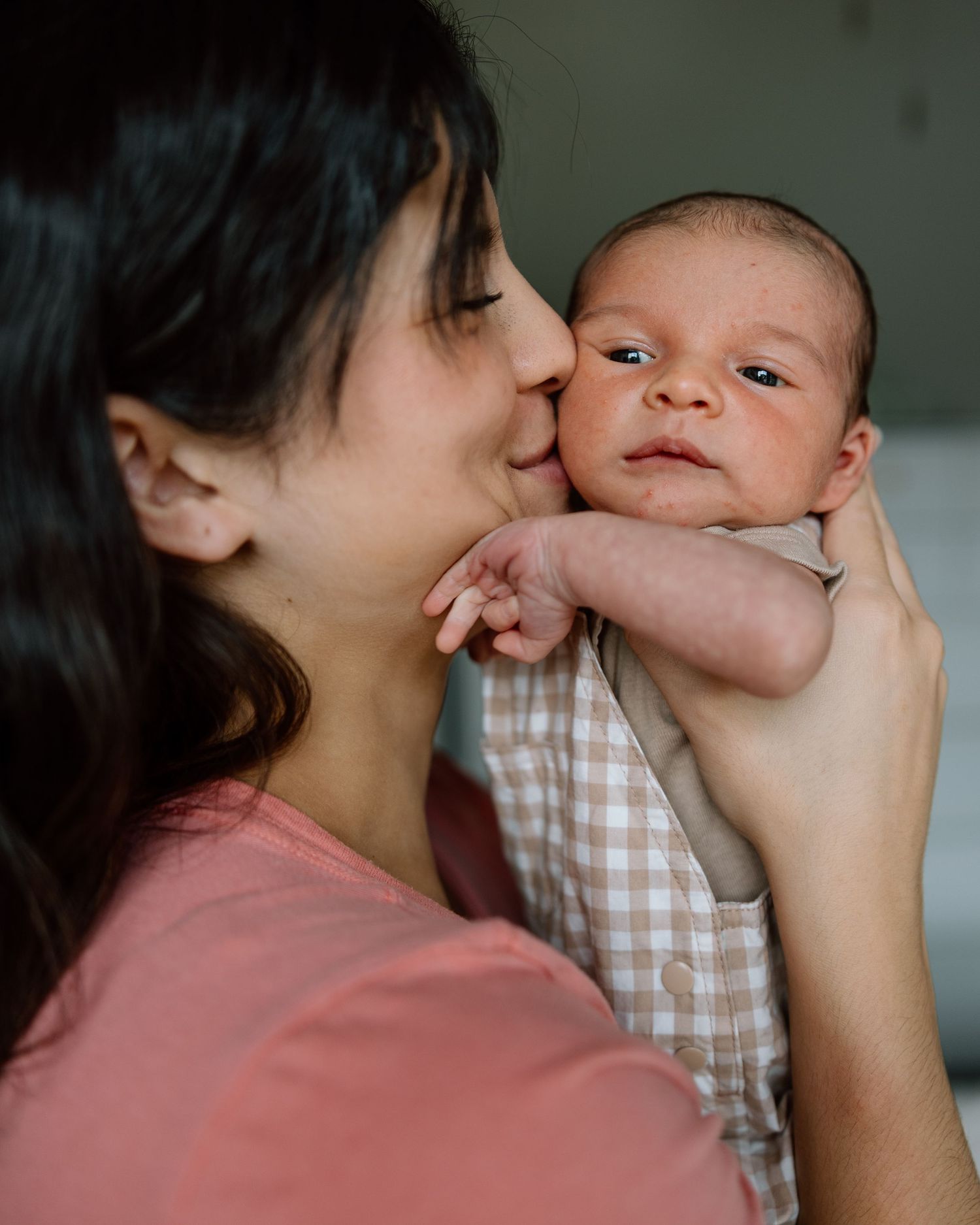New mother in pink shirt kissing her alert newborn babys cheek in a tender moment.