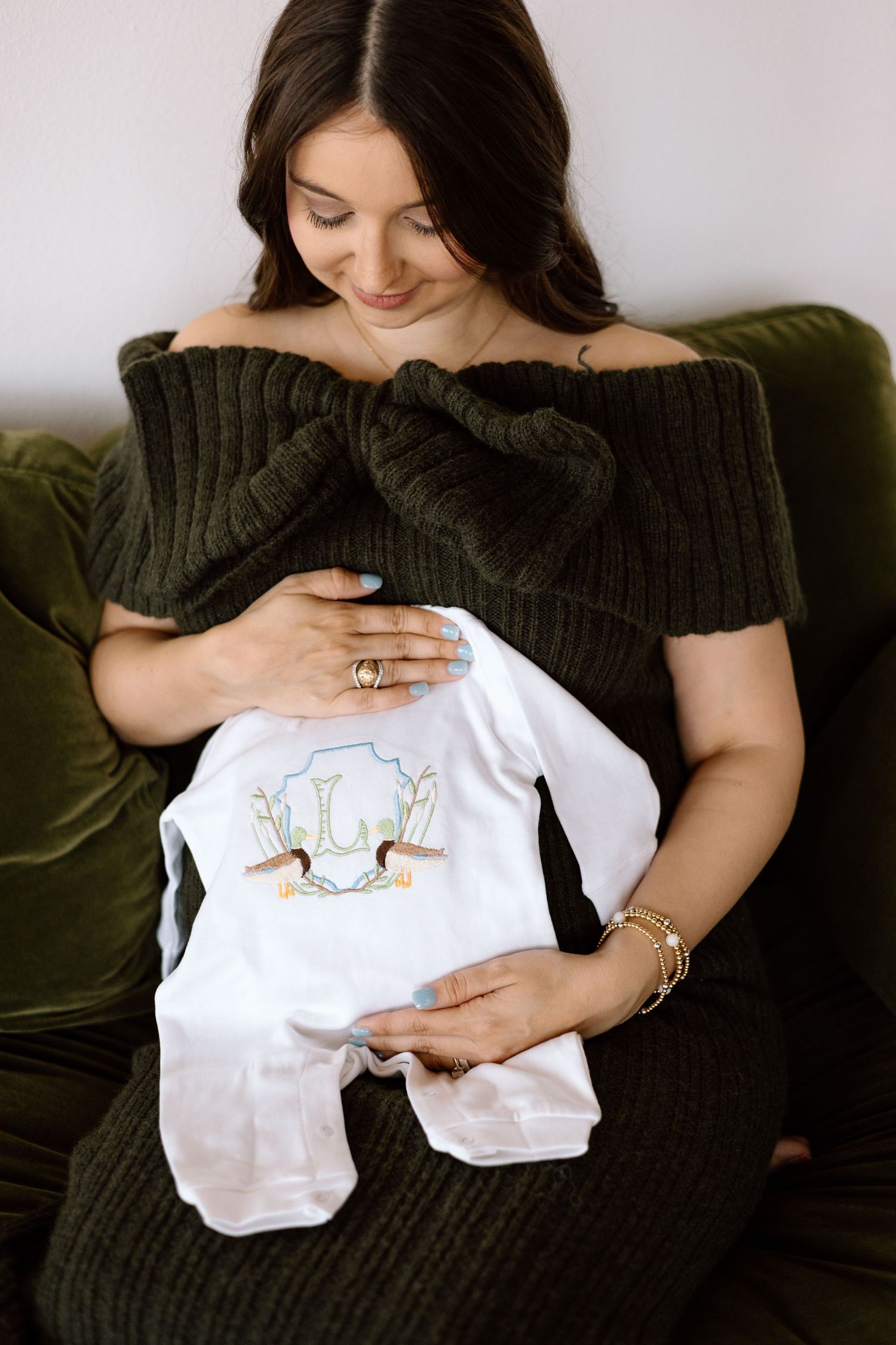 Pregnant brunette woman in dark green off-shoulder sweater dress holding baby onesie while seated on velvet sofa.