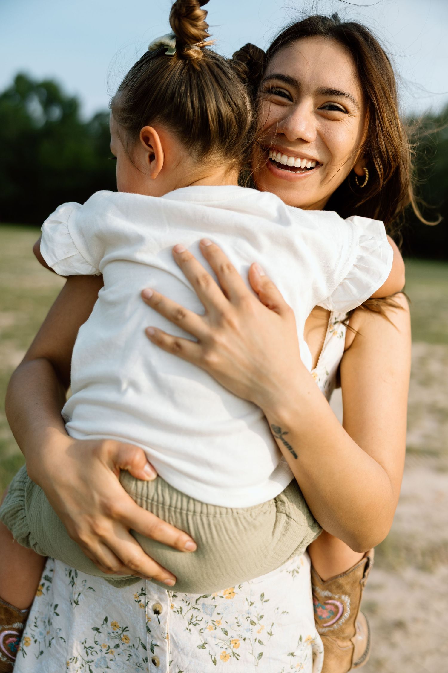 A joyful mother laughs while hugging her young daughter outdoors in a sunny field.