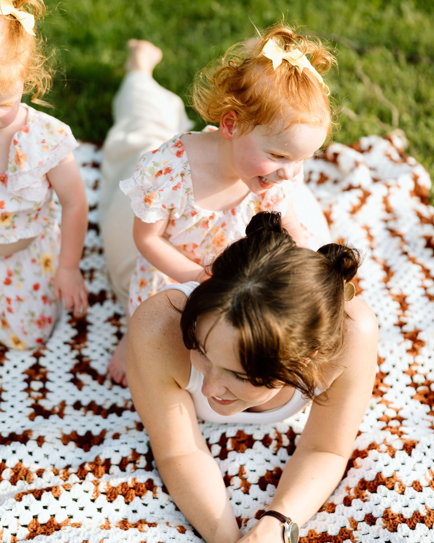 A mother plays with two red-haired toddler girls in floral dresses on a crocheted blanket outdoors.